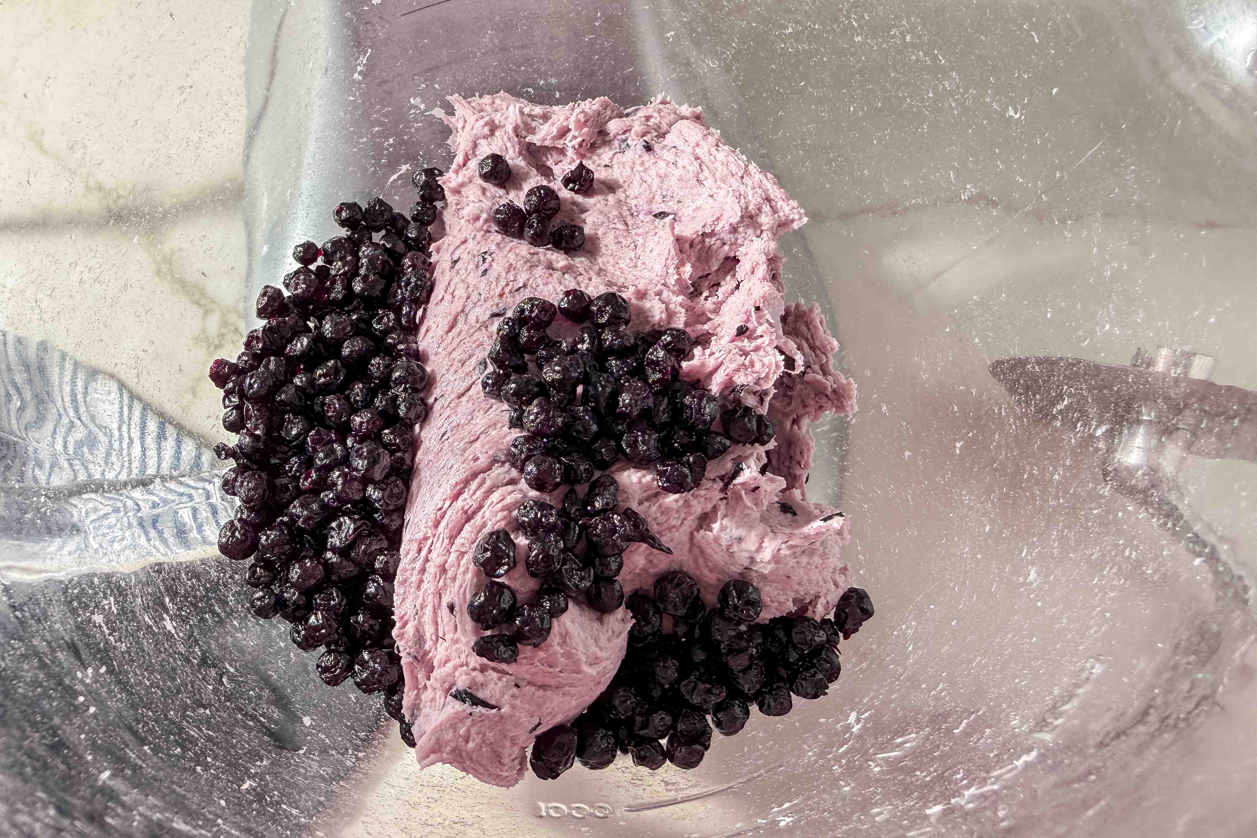 Purple blueberry bagel dough and whole dried blueberries sit on the side of a metal mixing bowl, with part of a blue and white striped cloth visible in the background.