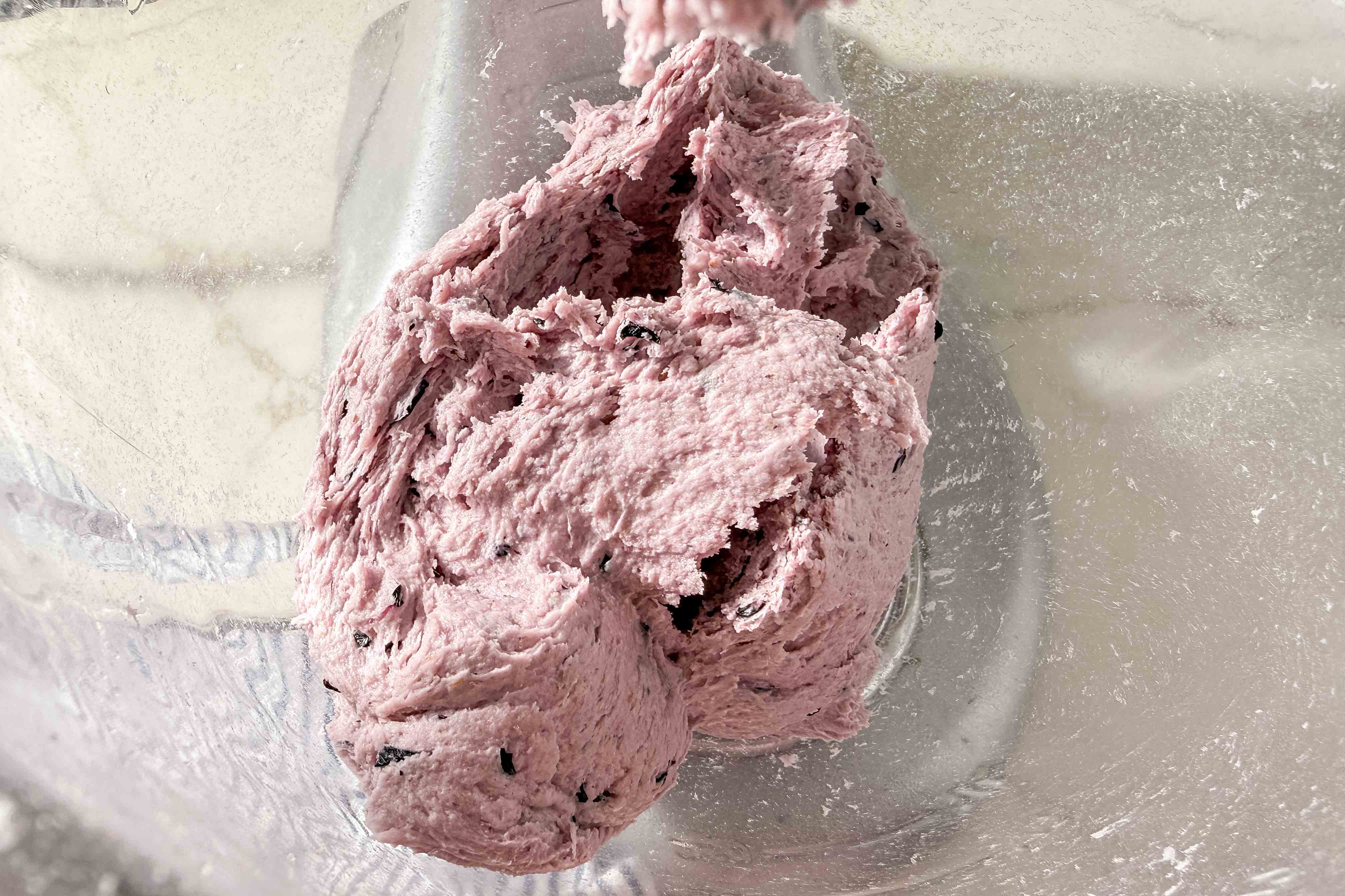 A close-up of a metal mixing bowl with purple bagel dough that contains dried blueberries sticking to the sides and the mixer attachment.
