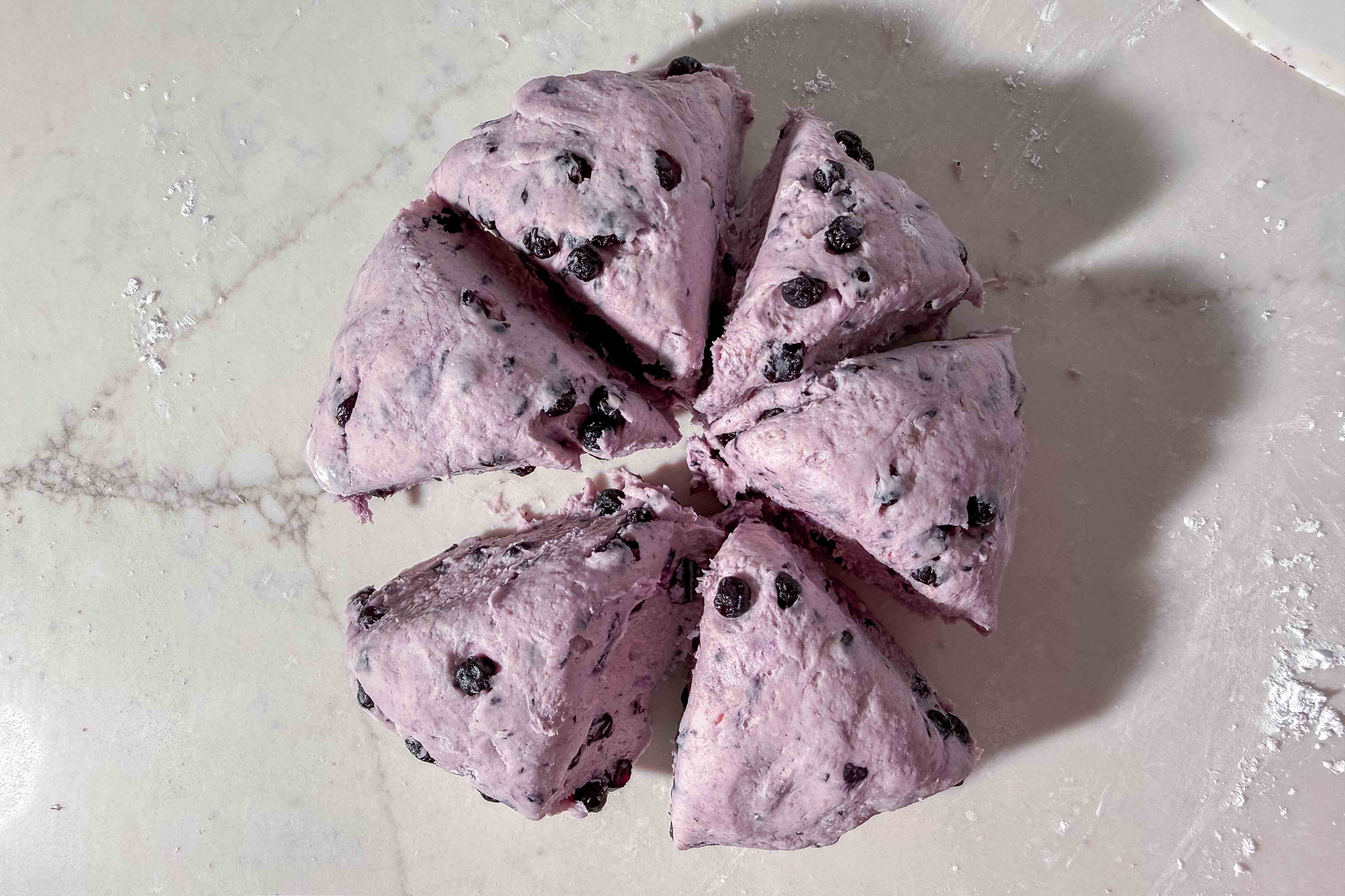Purple blueberry bagel dough cut into six triangular wedges, arranged in a circular pattern on a lightly floured white surface.