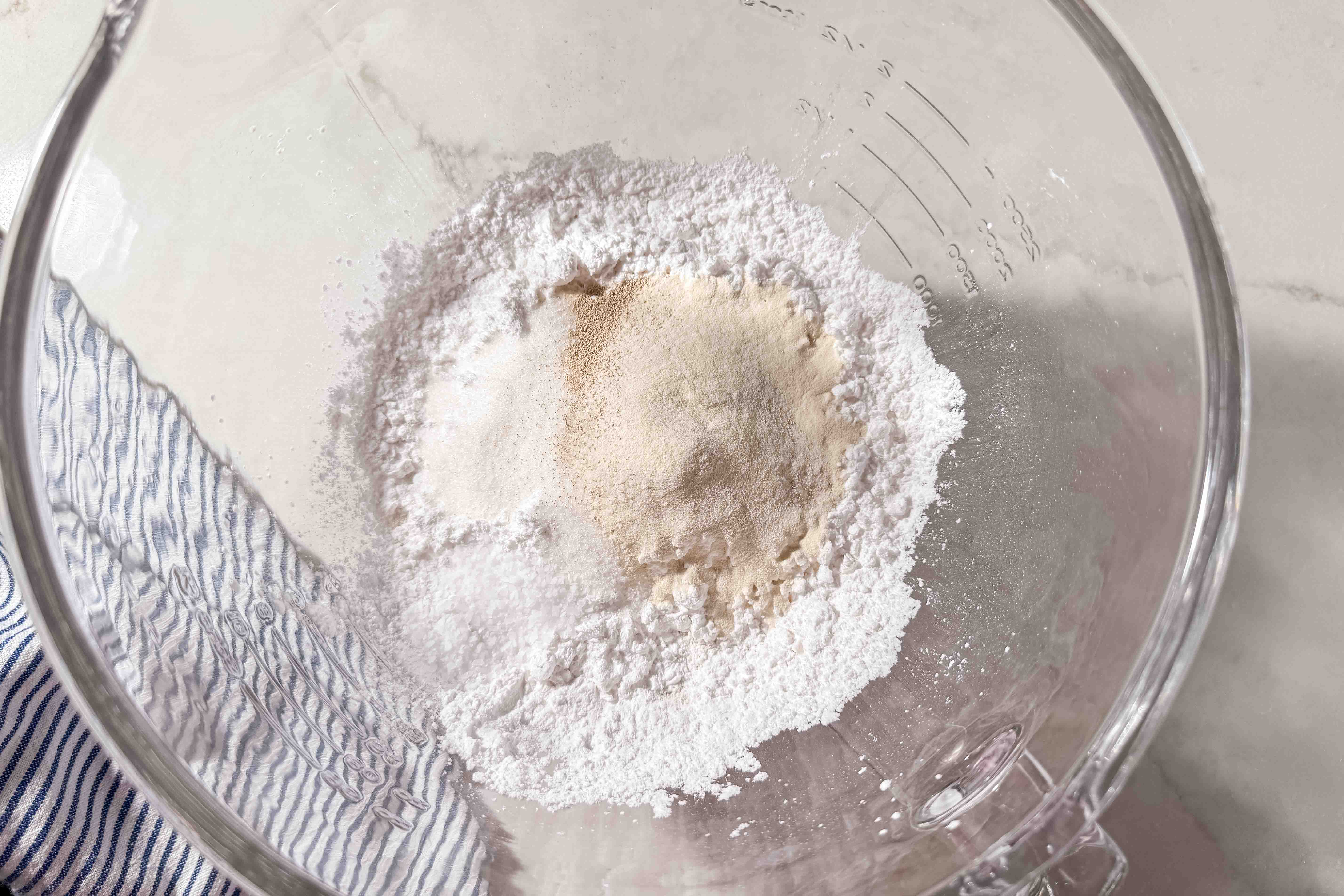A glass mixing bowl holds a mound of gluten-free flours and a small pile of yeast. A blue-and-white striped kitchen towel is next to the bowl on a light countertop.