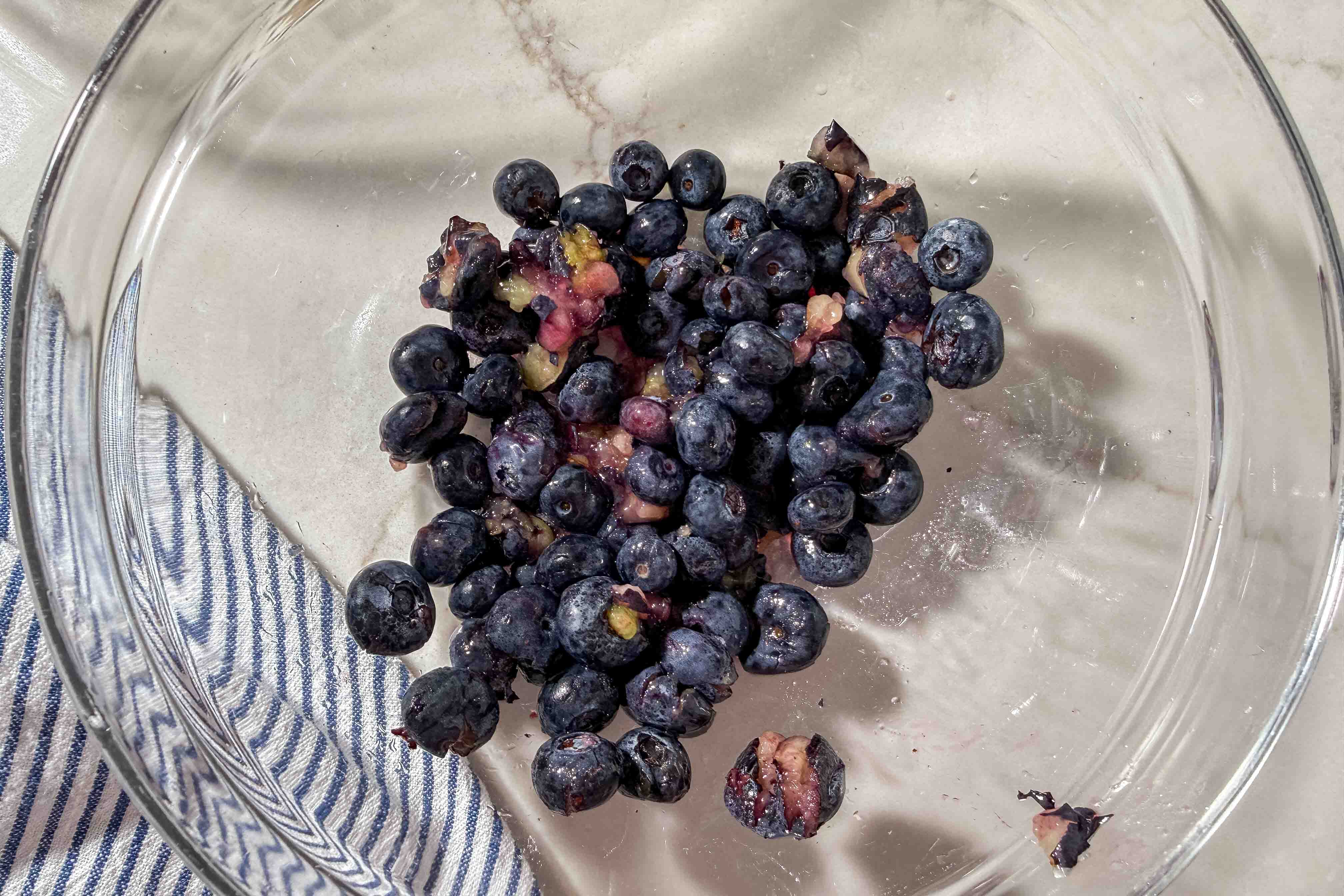 A glass bowl holds several slightly crushed blueberries, some with their juice leaking, on a striped blue and white cloth on a marble surface.