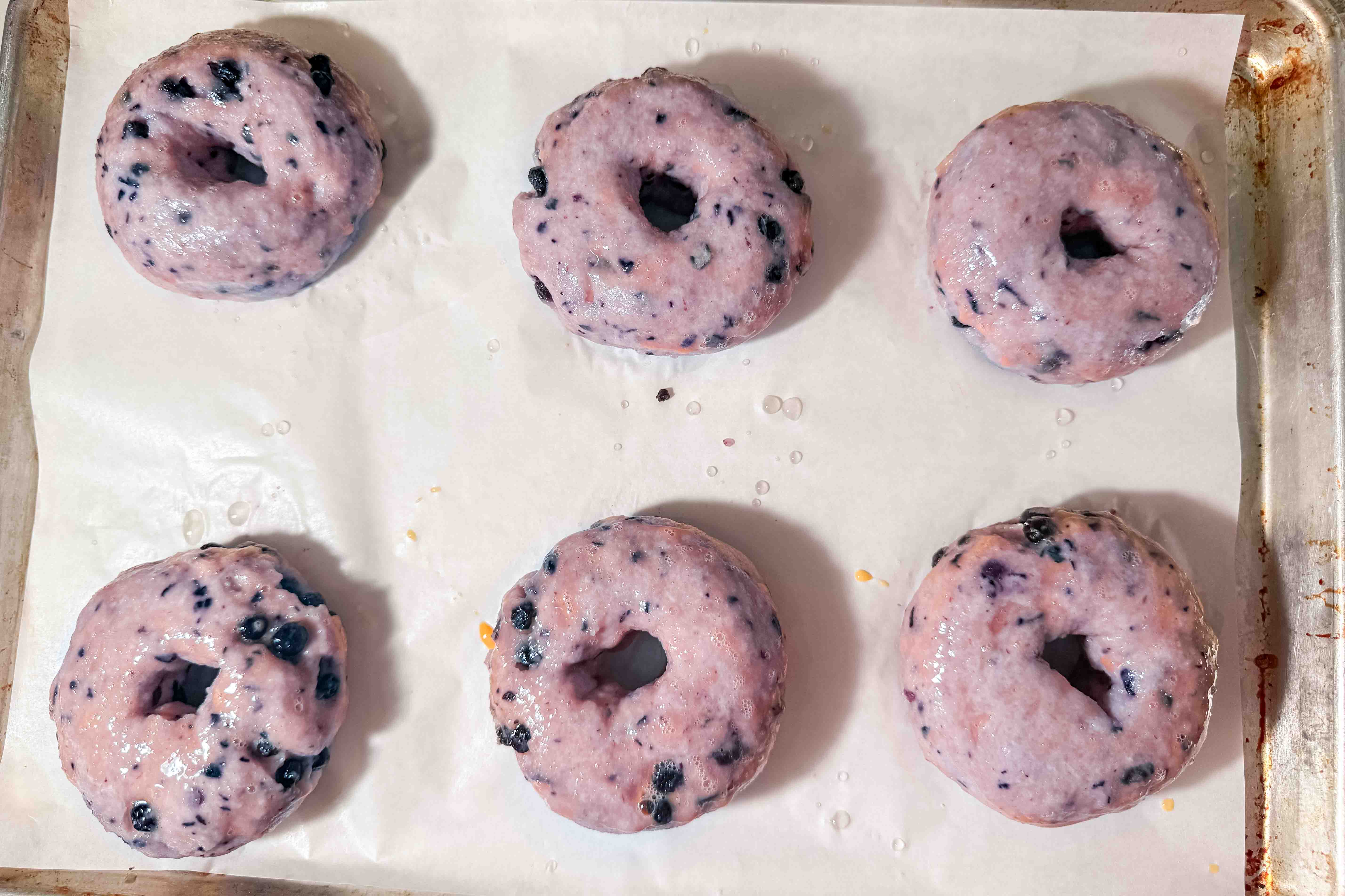 Six unbaked blueberry donuts with a light purple hue rest on a baking sheet lined with parchment paper.