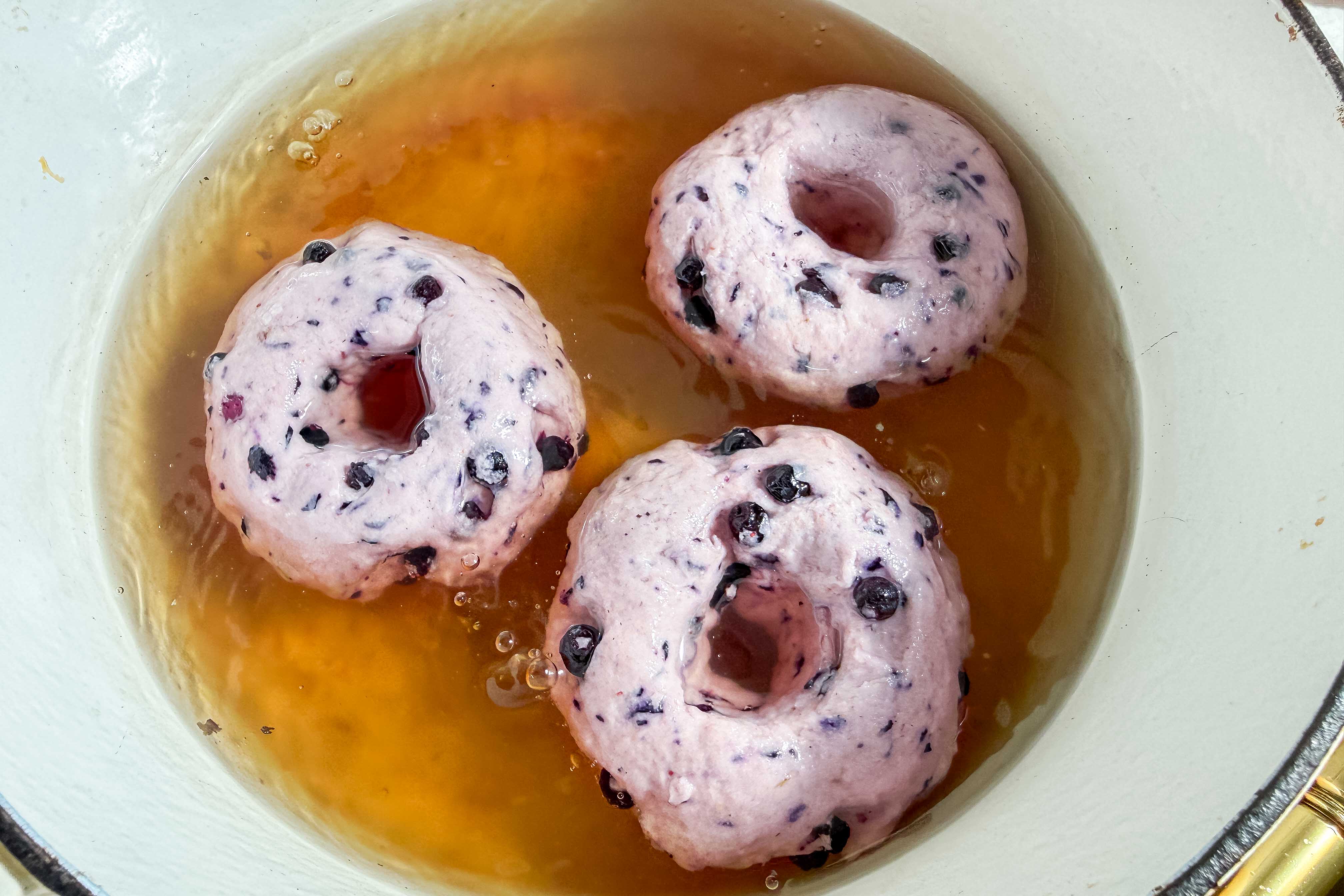 Three uncooked gluten-free blueberry donuts are boiling in a water and brown sugar mixture inside a white pot. The donuts are light purple with visible blueberries.