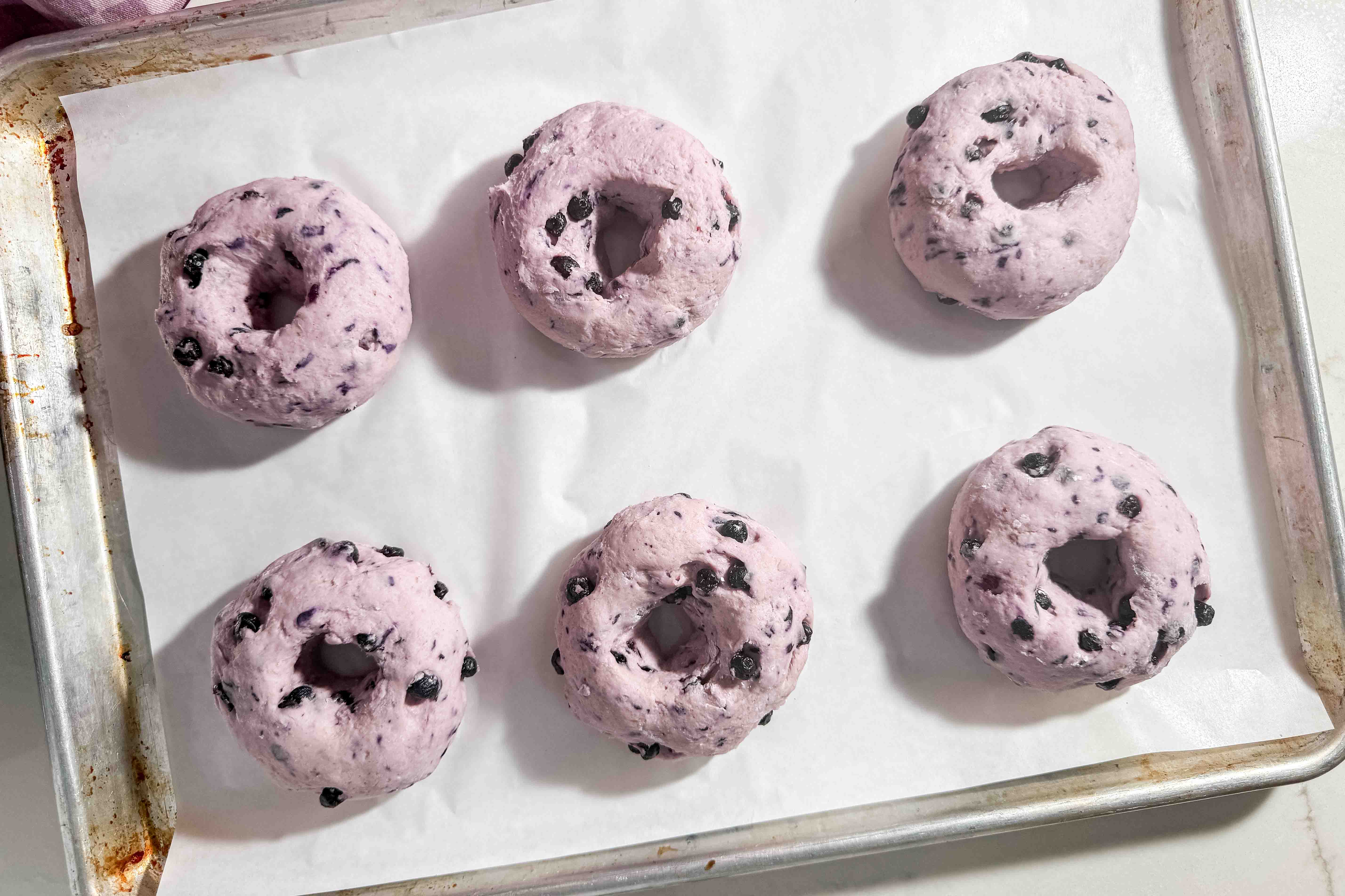 Six unbaked, purple blueberry bagels with visible blueberries are arranged on a parchment-lined baking sheet, ready to be baked.