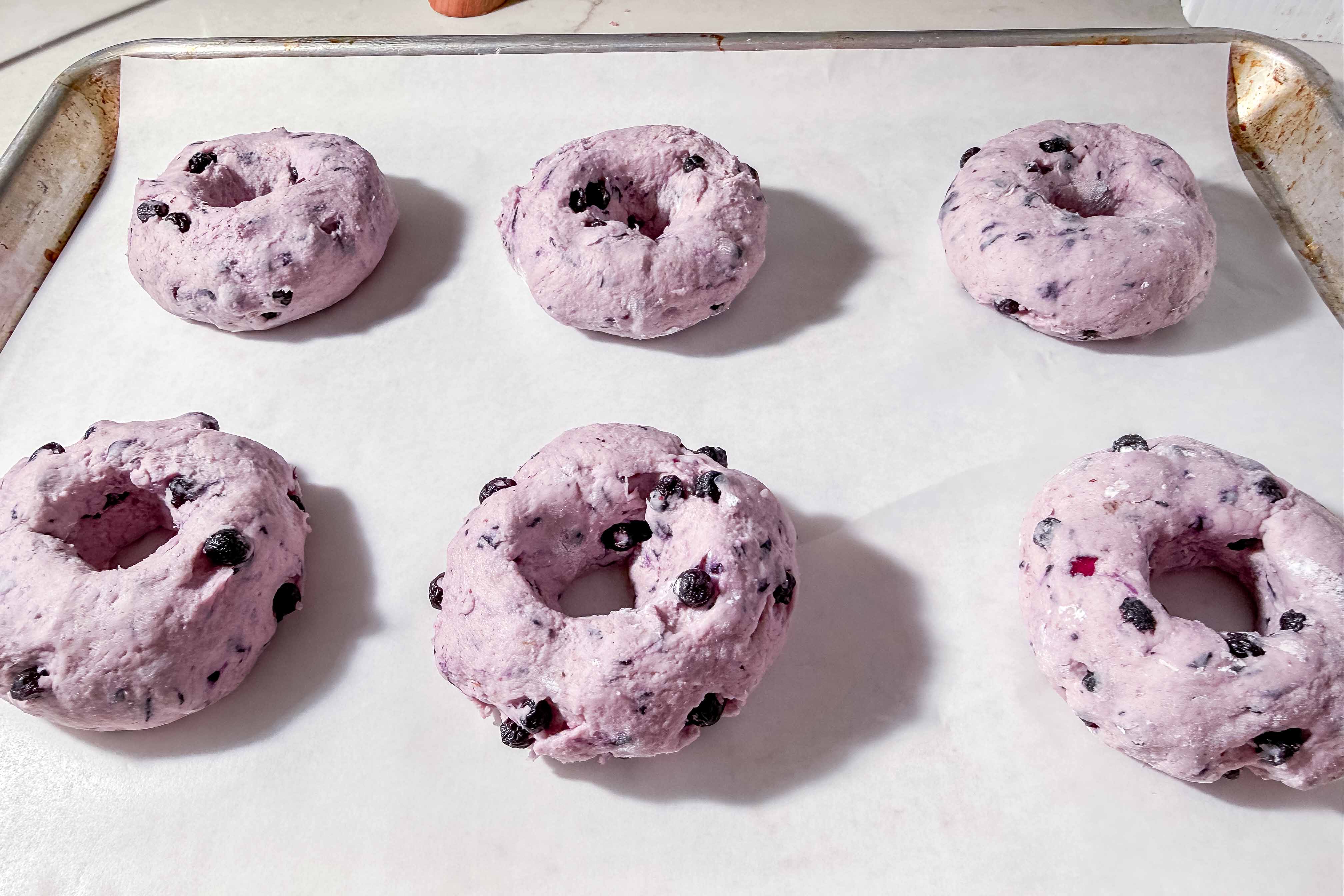 Six unbaked, purple blueberry bagels on a parchment-lined baking sheet, ready to go into the oven.