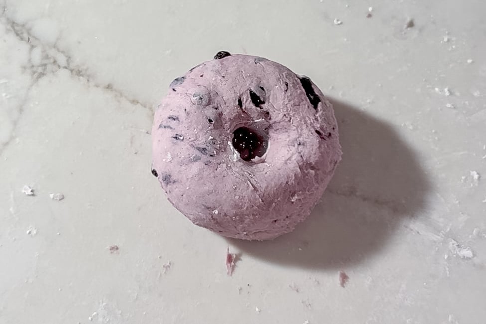 A round, purple dough ball with dried blueberries, sits on a white marble surface. The dough has a small hole in the center.