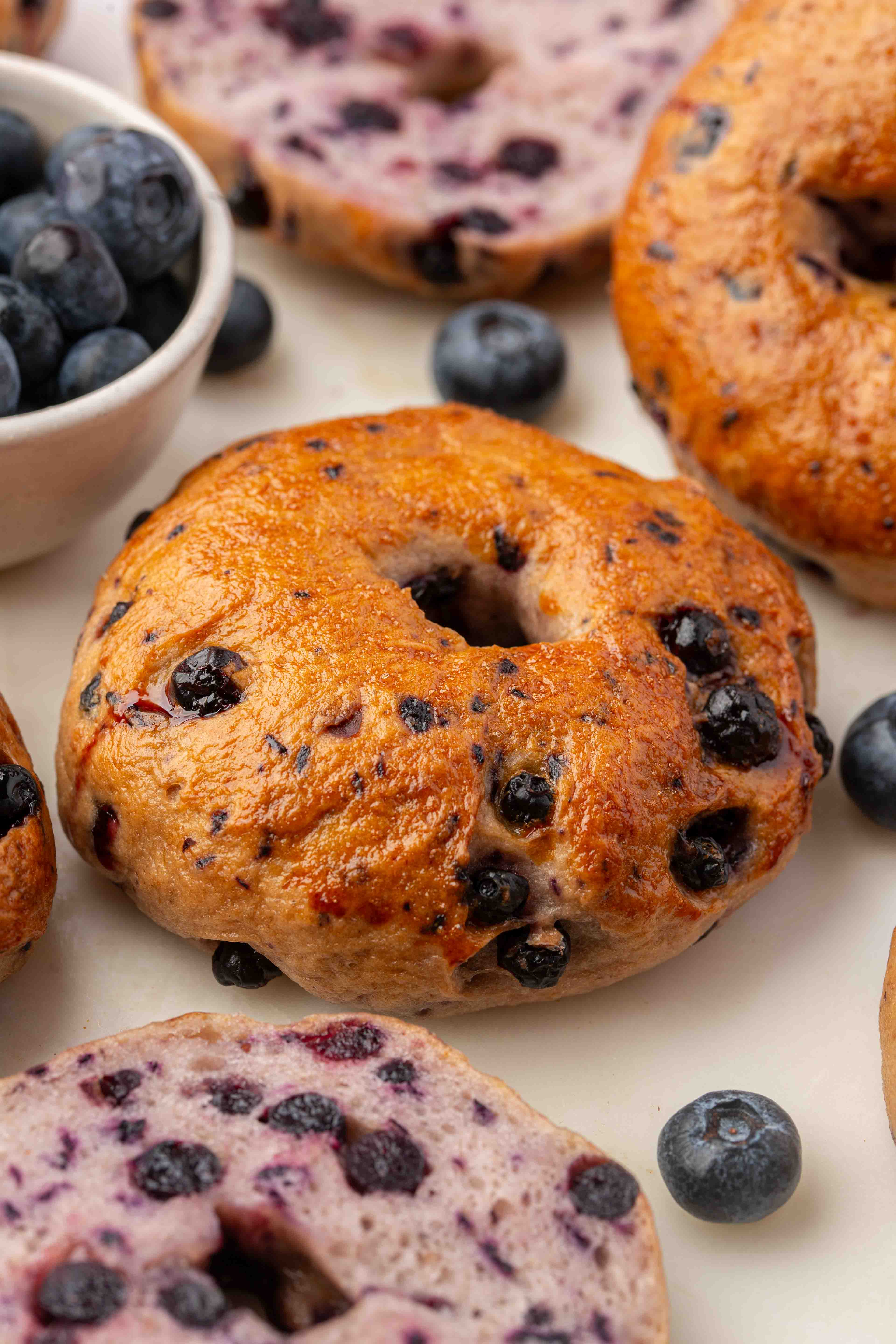 Close-up of blueberry bagels, some whole and some sliced, surrounded by fresh blueberries and a small bowl of blueberries on a white surface.