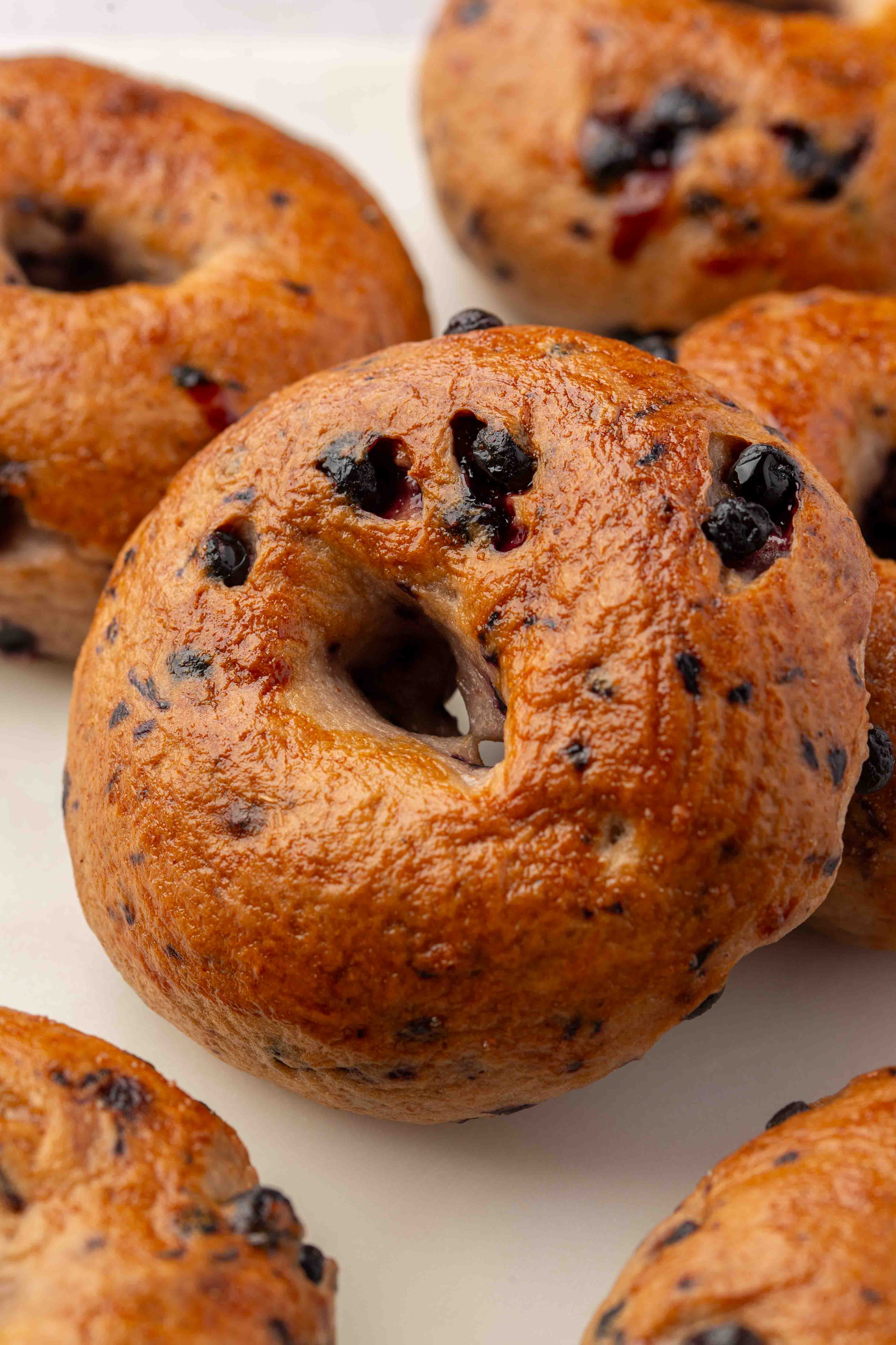 Close-up of several golden-brown gluten-free blueberry bagels on a white surface, showing their shiny, textured crusts and scattered baked-in blueberries.