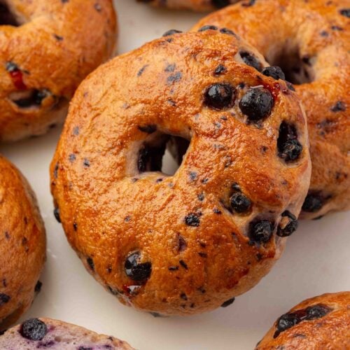 A close-up of several blueberry bagels on a white surface, showing golden-brown crusts and visible blueberries baked into the dough.