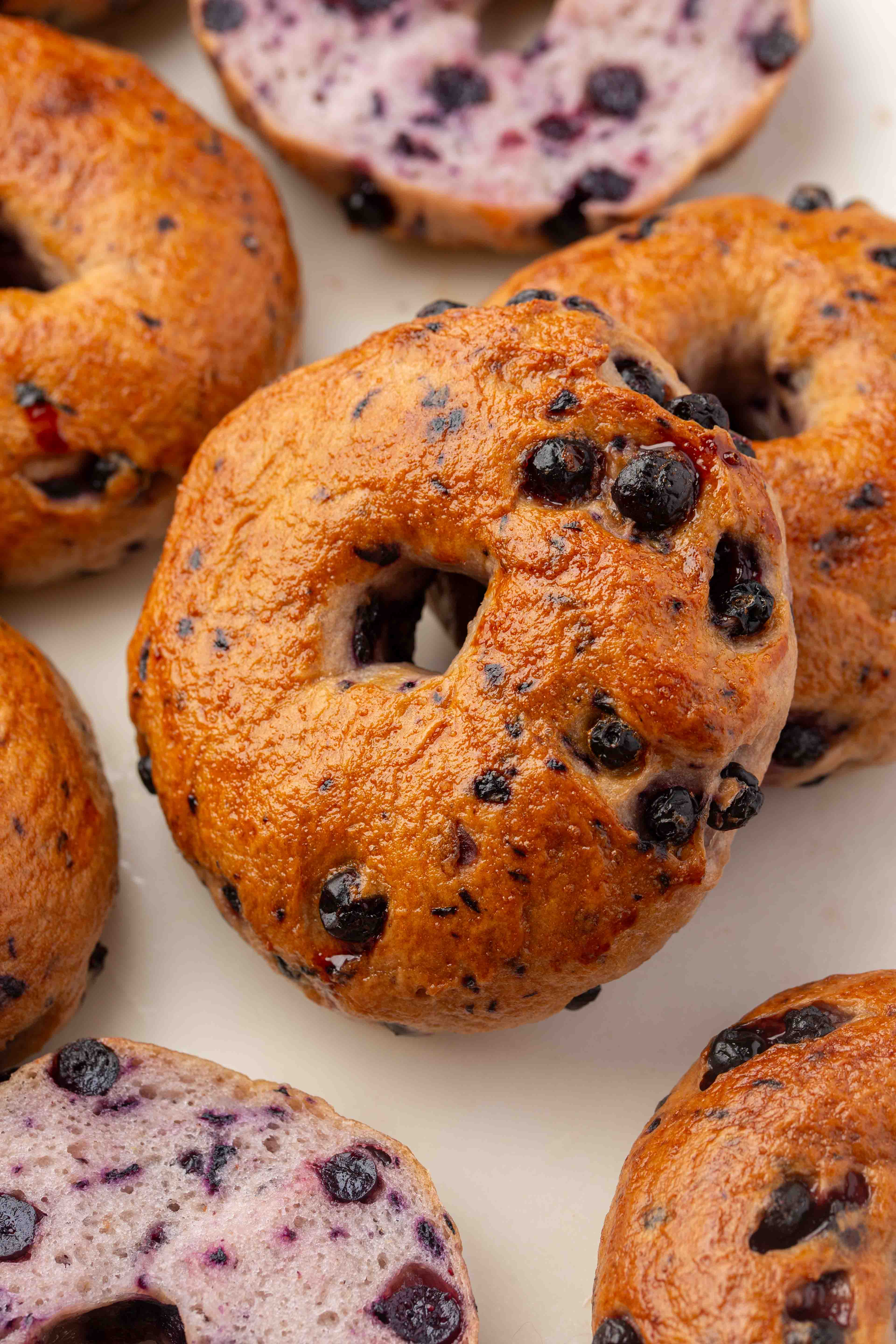A close-up of several blueberry bagels on a white surface, showing golden-brown crusts and visible blueberries baked into the dough.