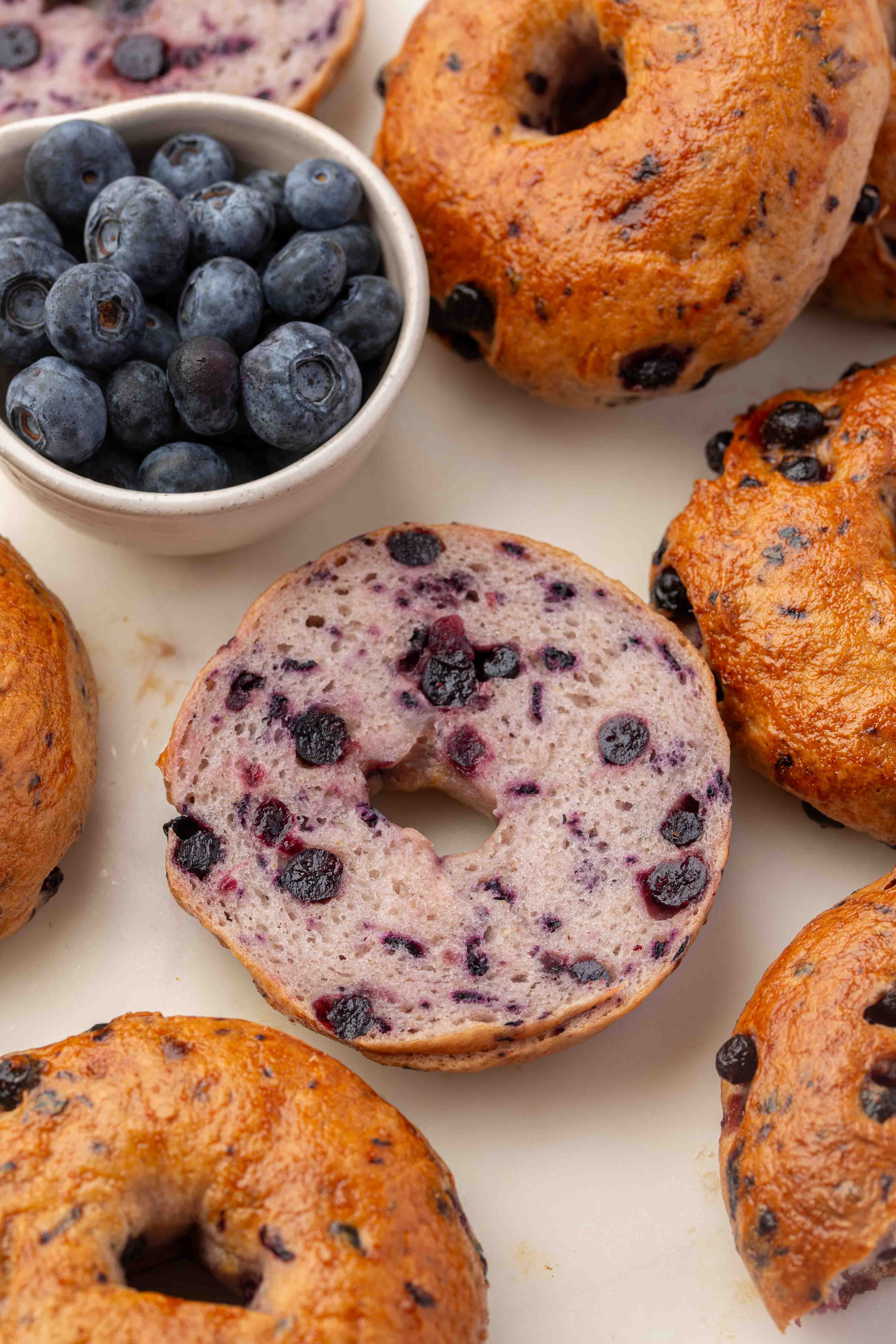 A sliced blueberry bagel sits among whole blueberry bagels, with a small bowl of fresh blueberries nearby on a light surface.