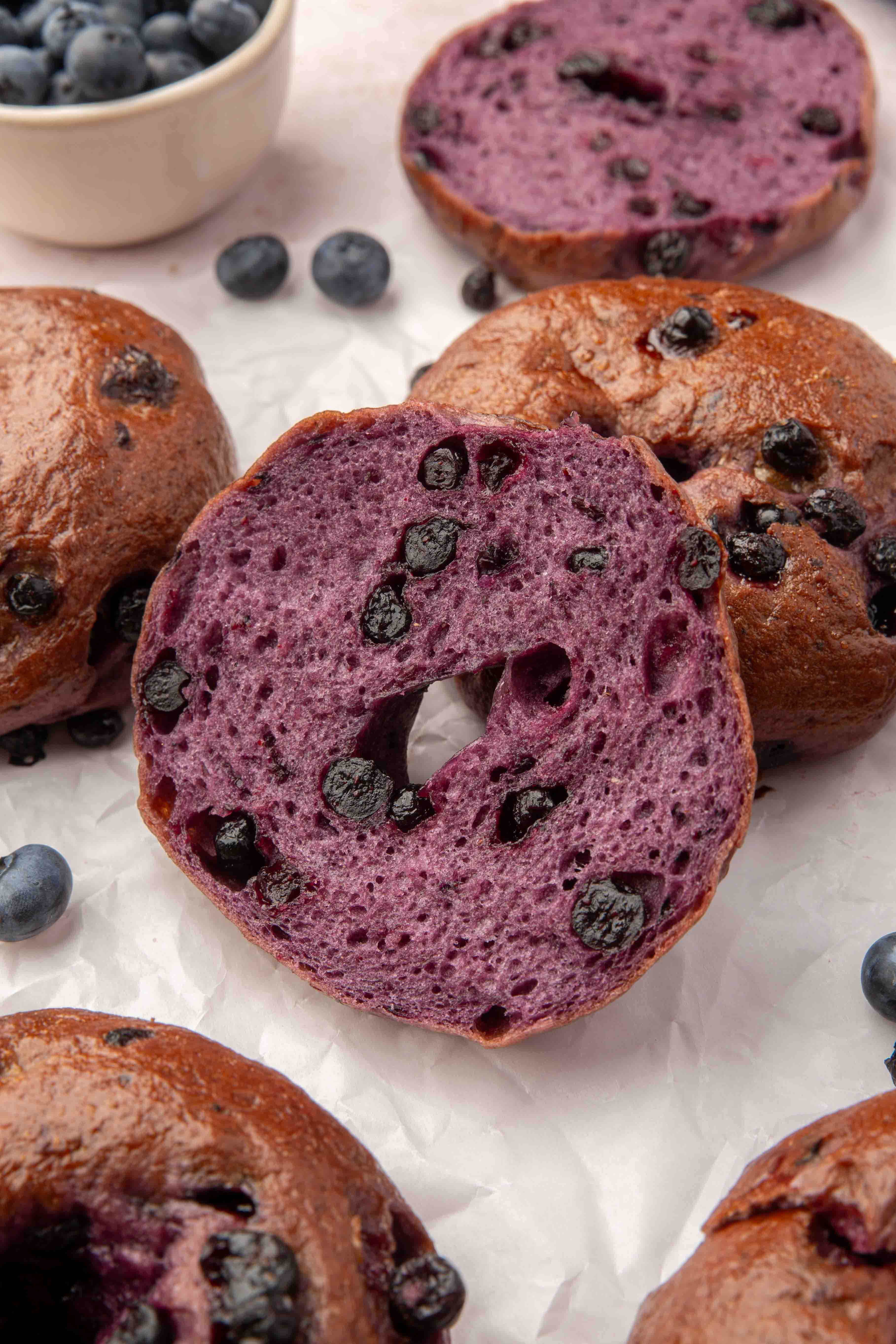 A sliced blueberry bagel with a vibrant purple interior and visible blueberries, surrounded by whole bagels and fresh blueberries on a white surface.