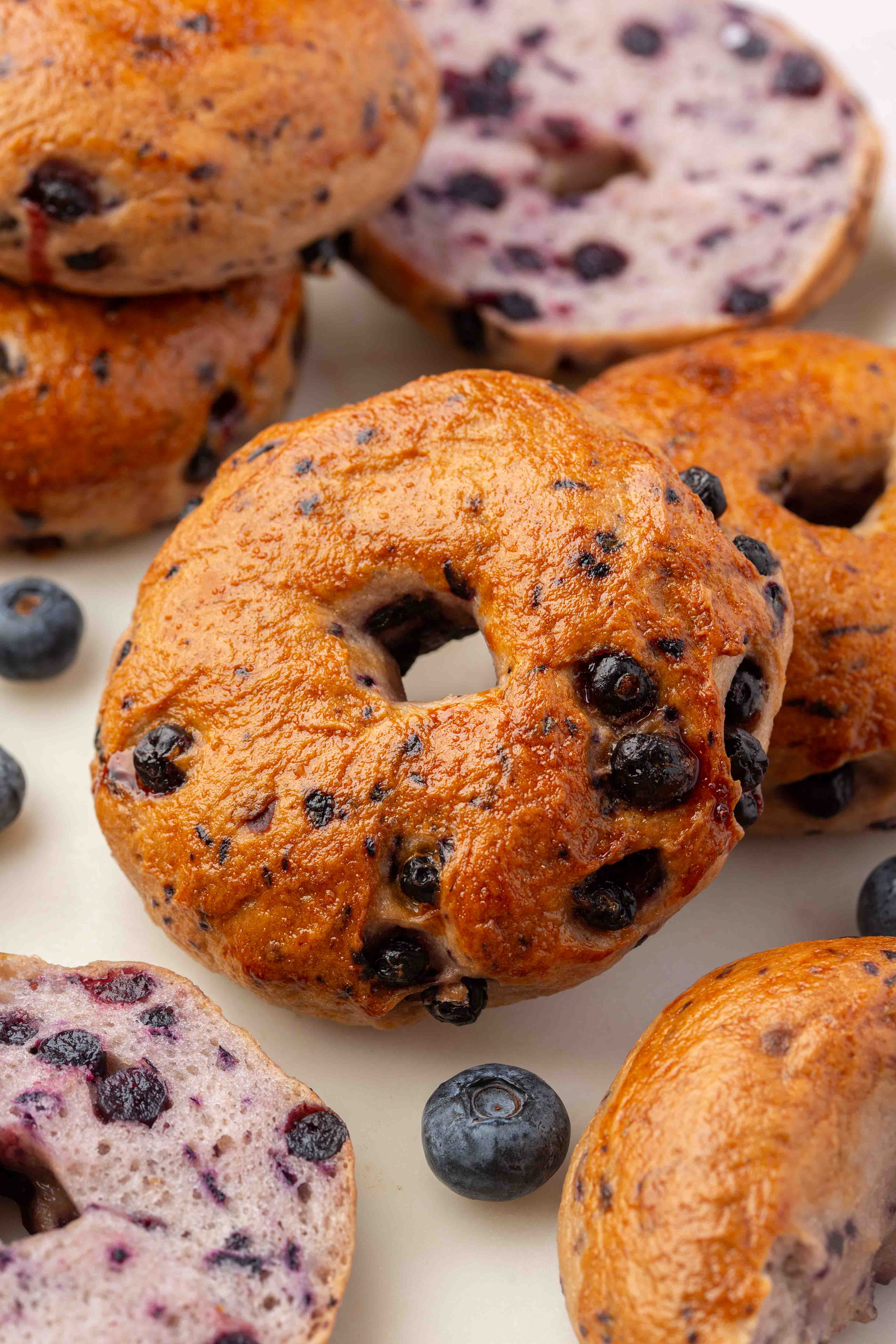 Blueberry bagels, some whole and some sliced, are arranged on a white surface with a few loose blueberries scattered around.