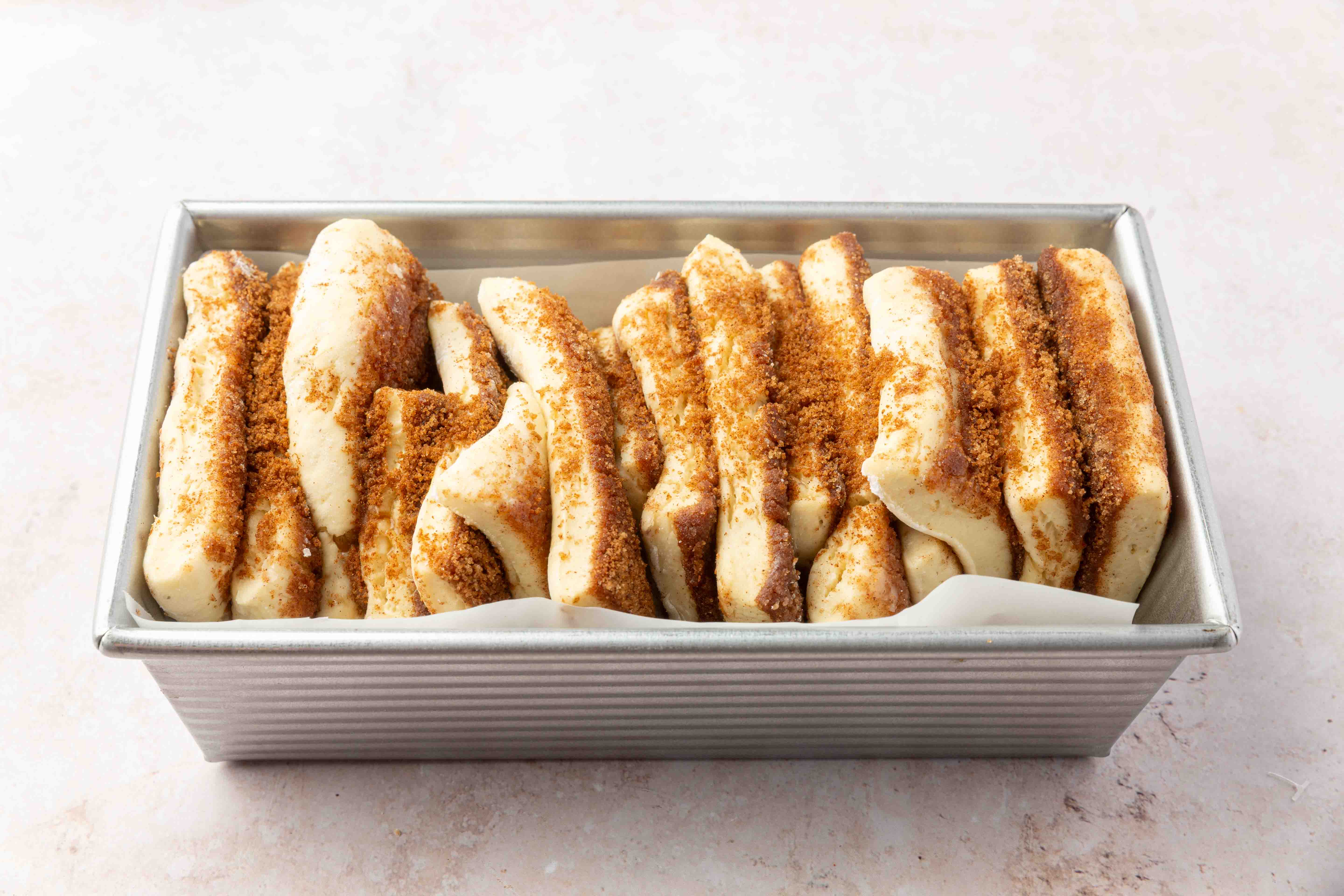 A loaf pan filled with layers of uncooked cinnamon pull-apart bread dough, sprinkled with brown sugar and cinnamon, ready to be baked.