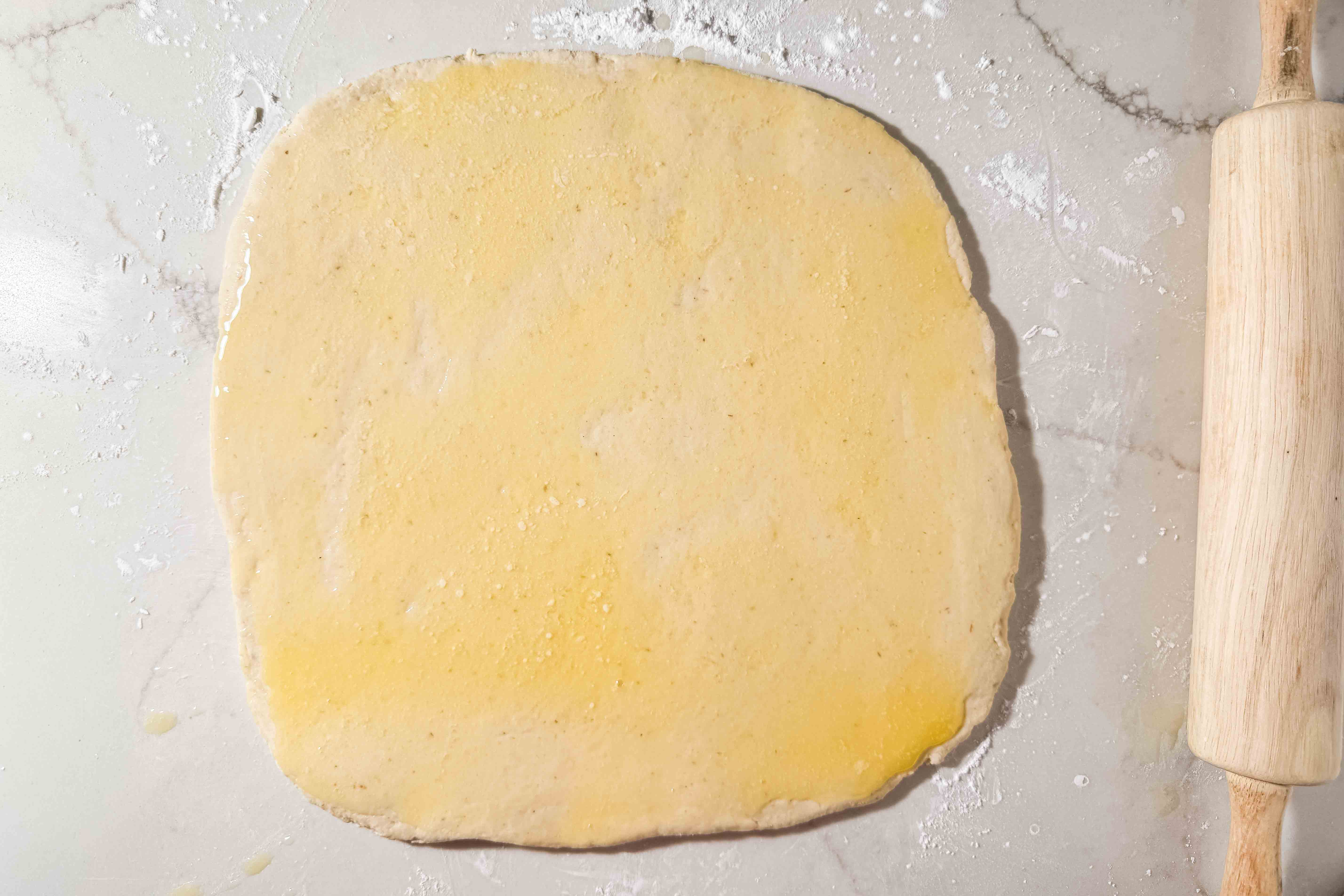 A square of rolled-out dough brushed with melted butter sits on a floured surface next to a wooden rolling pin.