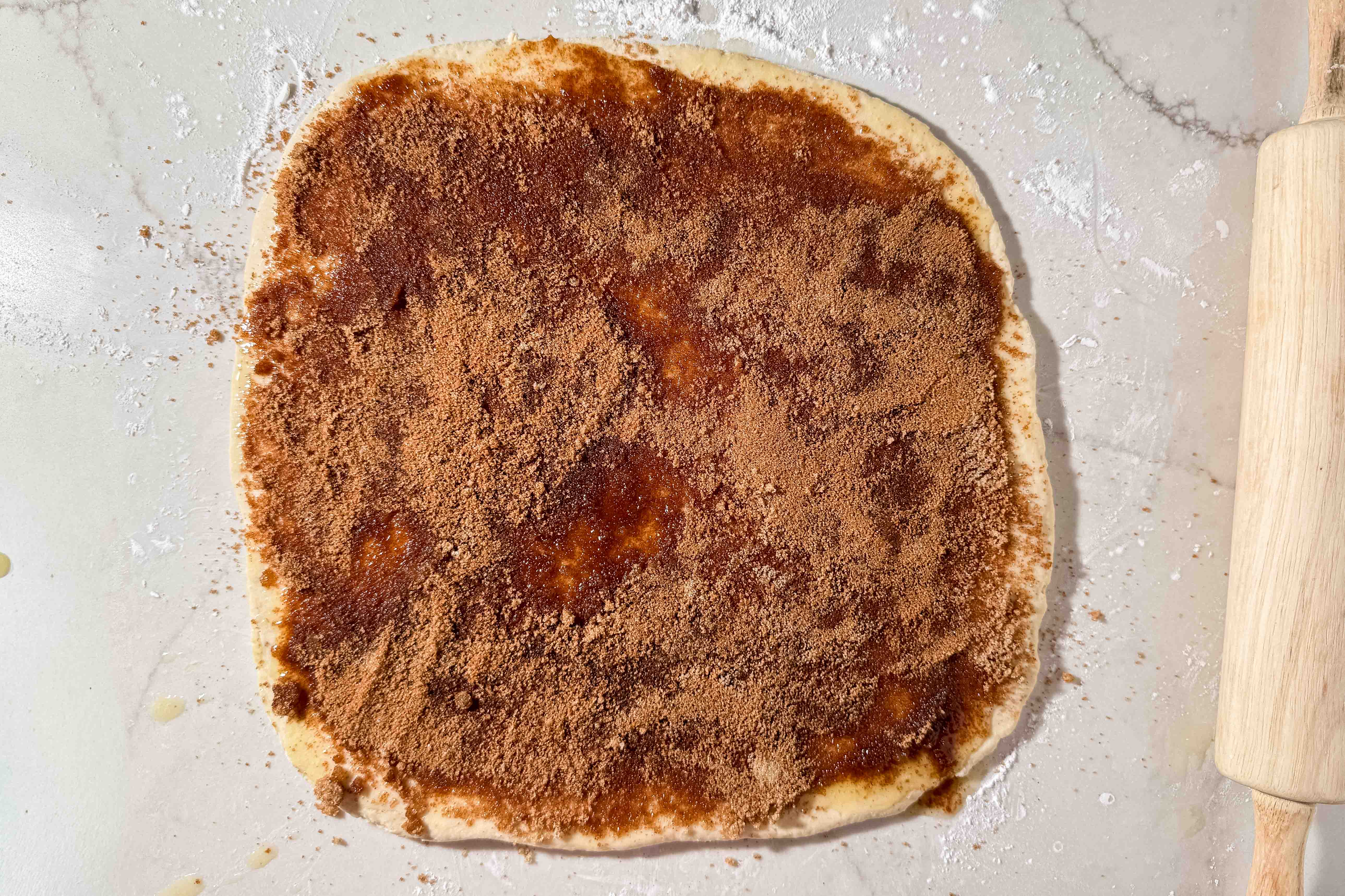 A square sheet of dough covered with a brown cinnamon-sugar mixture sits on a floured surface next to a rolling pin.