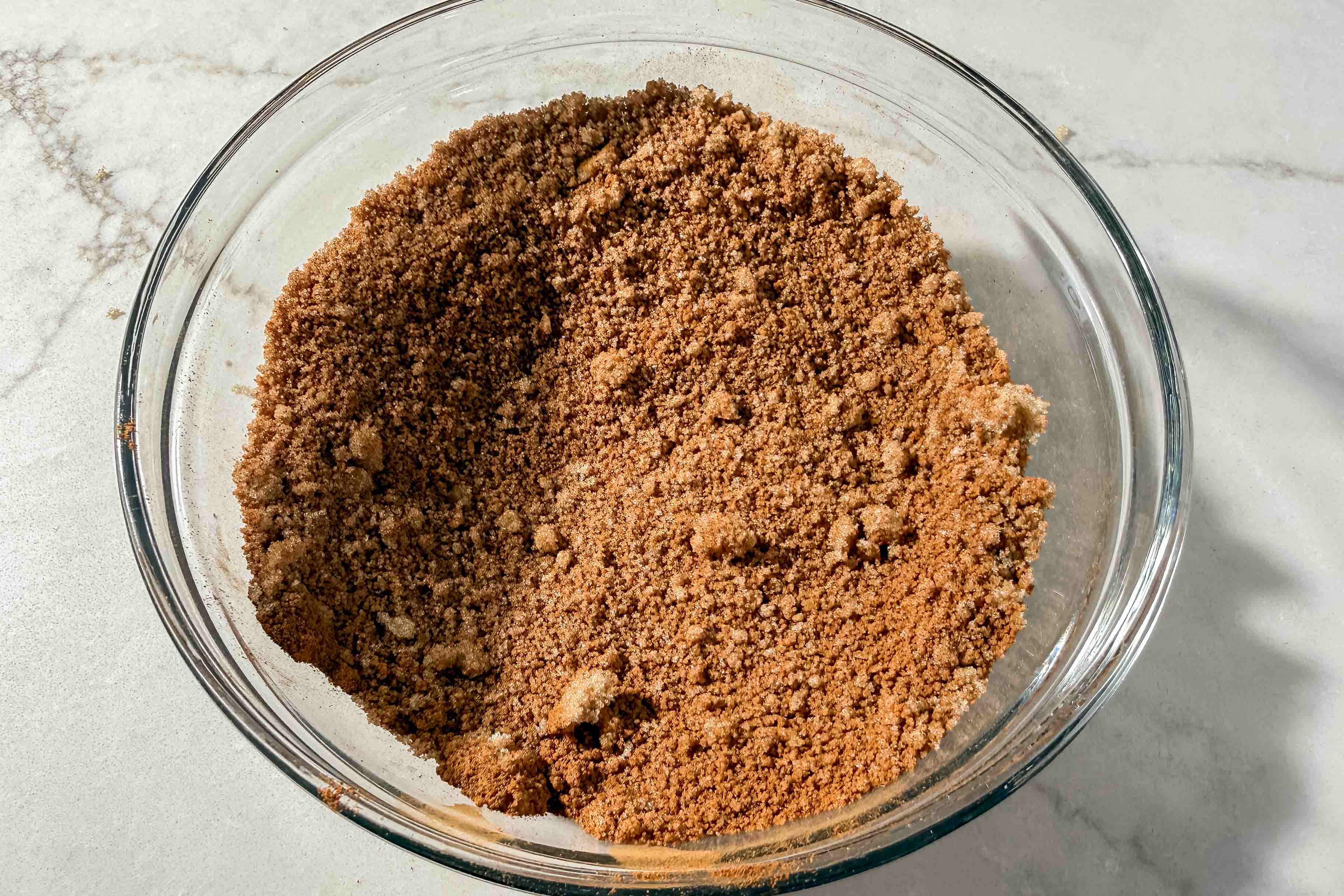 A clear glass bowl filled with brown sugar and cinnamon mixture sits on a light marble countertop.