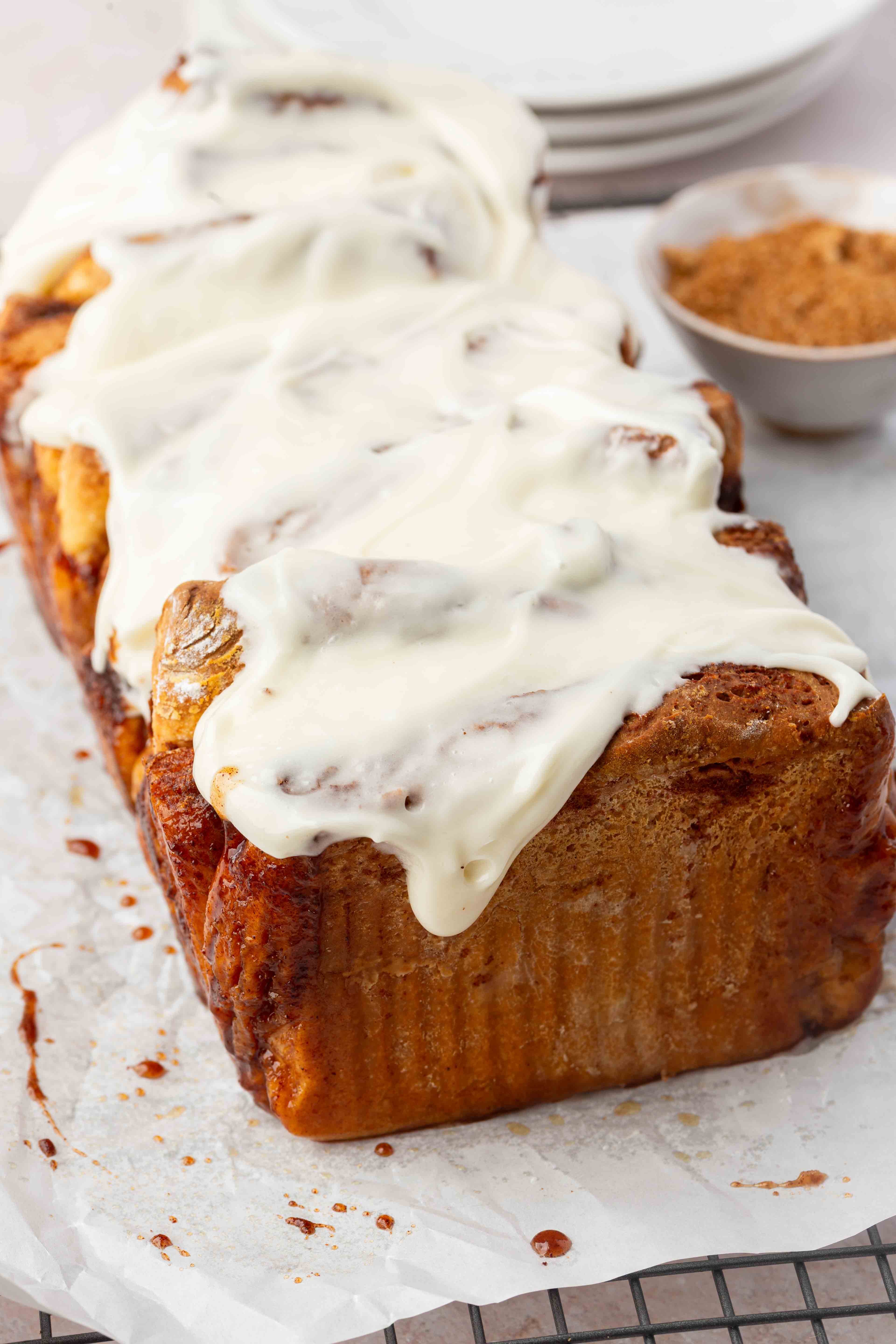 A loaf of pull-apart cinnamon bread topped with creamy white icing sits on a cooling rack, with a small bowl of ground cinnamon and stacked white plates in the background.