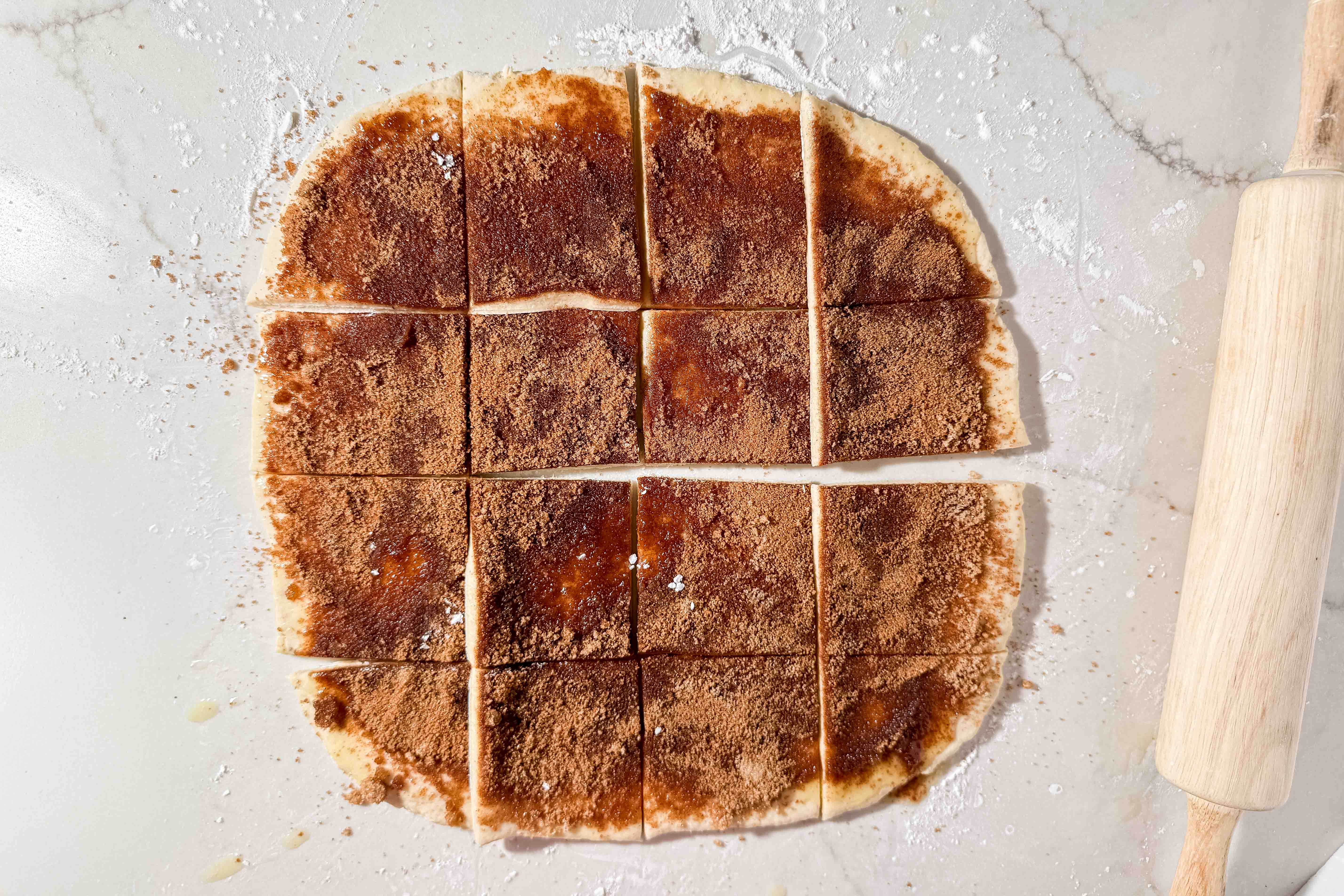 Dough rolled into a rectangle, topped with cinnamon sugar, and cut into 16 squares. A wooden rolling pin rests on the right side. Tapioca flour is scattered on the light countertop.