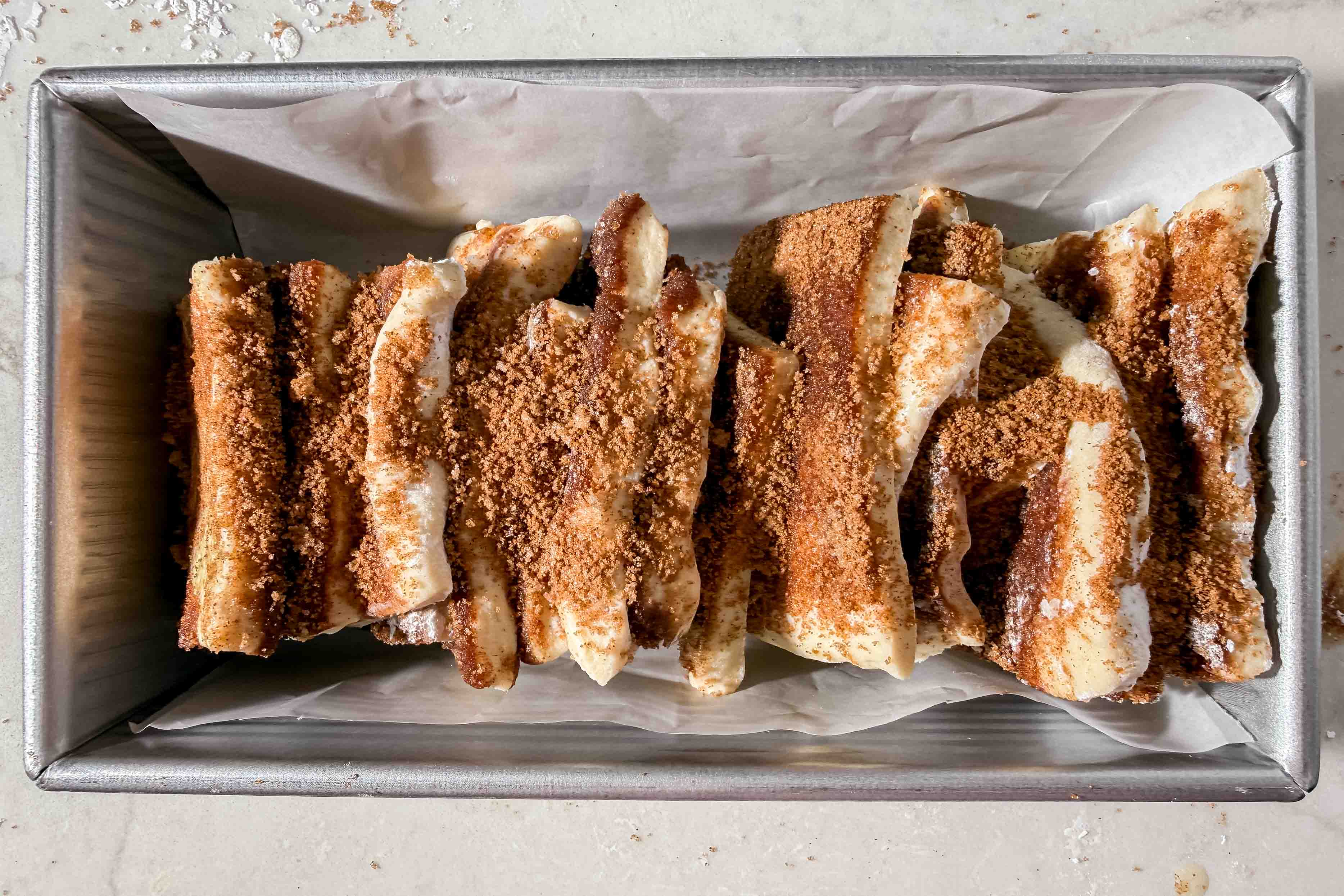 Slices of dough coated with cinnamon sugar are stacked in a parchment-lined loaf pan, ready for baking.