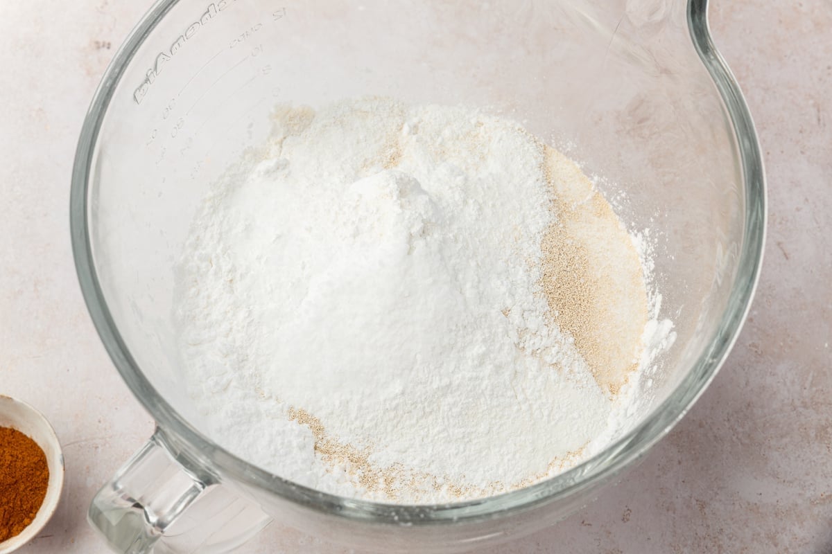 A glass bowl filled with dry baking ingredients, including tapioca flour, brown rice flour, potato starch, instant yeast, sugar, and baking powder, sits on a light countertop. A small dish with cinnamon is partially visible nearby.
