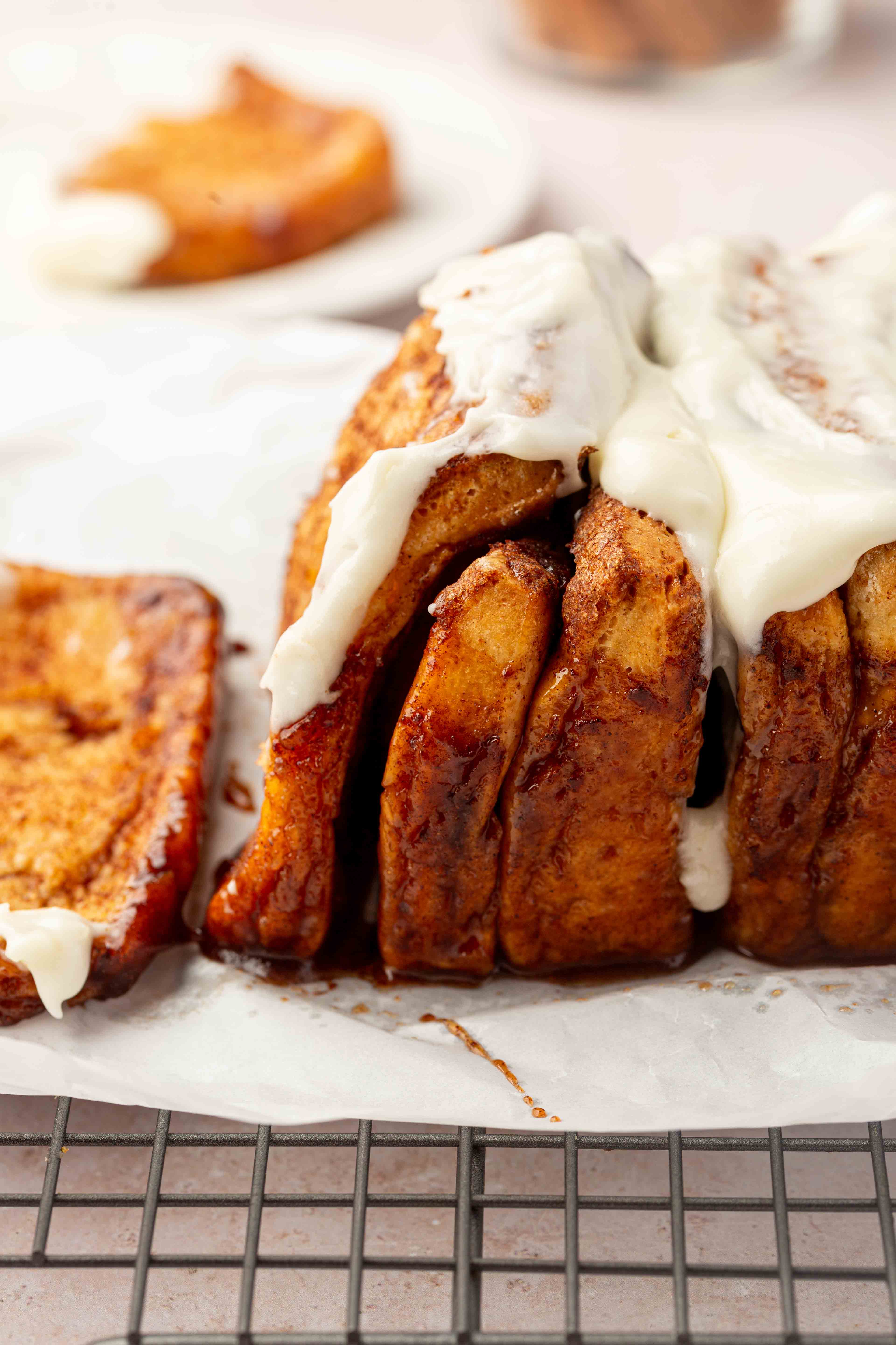 Close-up of cinnamon pull-apart bread topped with creamy white icing, sitting on parchment paper with a cooling rack underneath and a slice separated on the side.