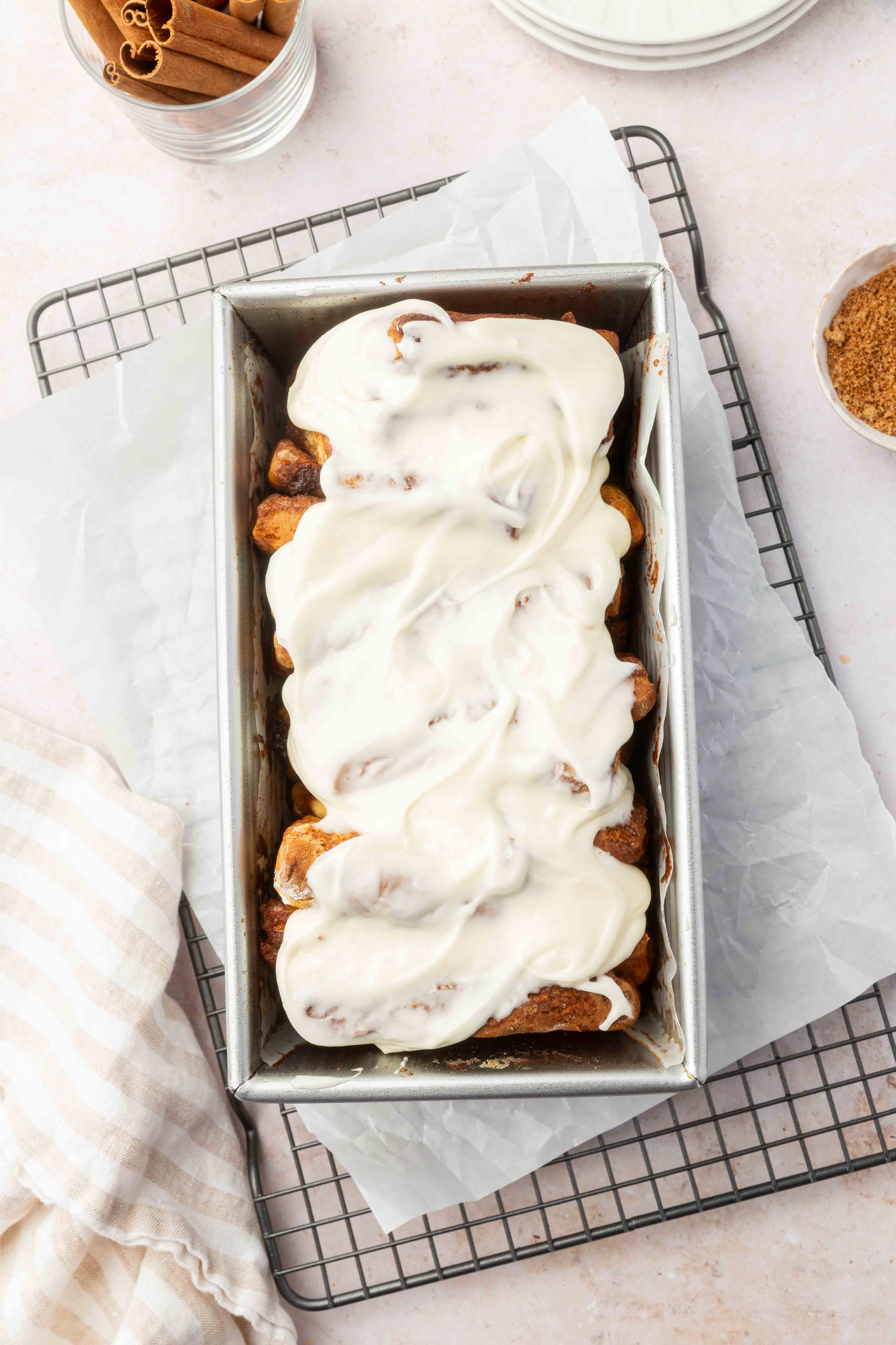 A loaf pan with cinnamon rolls topped with cream cheese icing sits on parchment paper and a cooling rack, surrounded by bowls of cinnamon sticks and brown sugar.