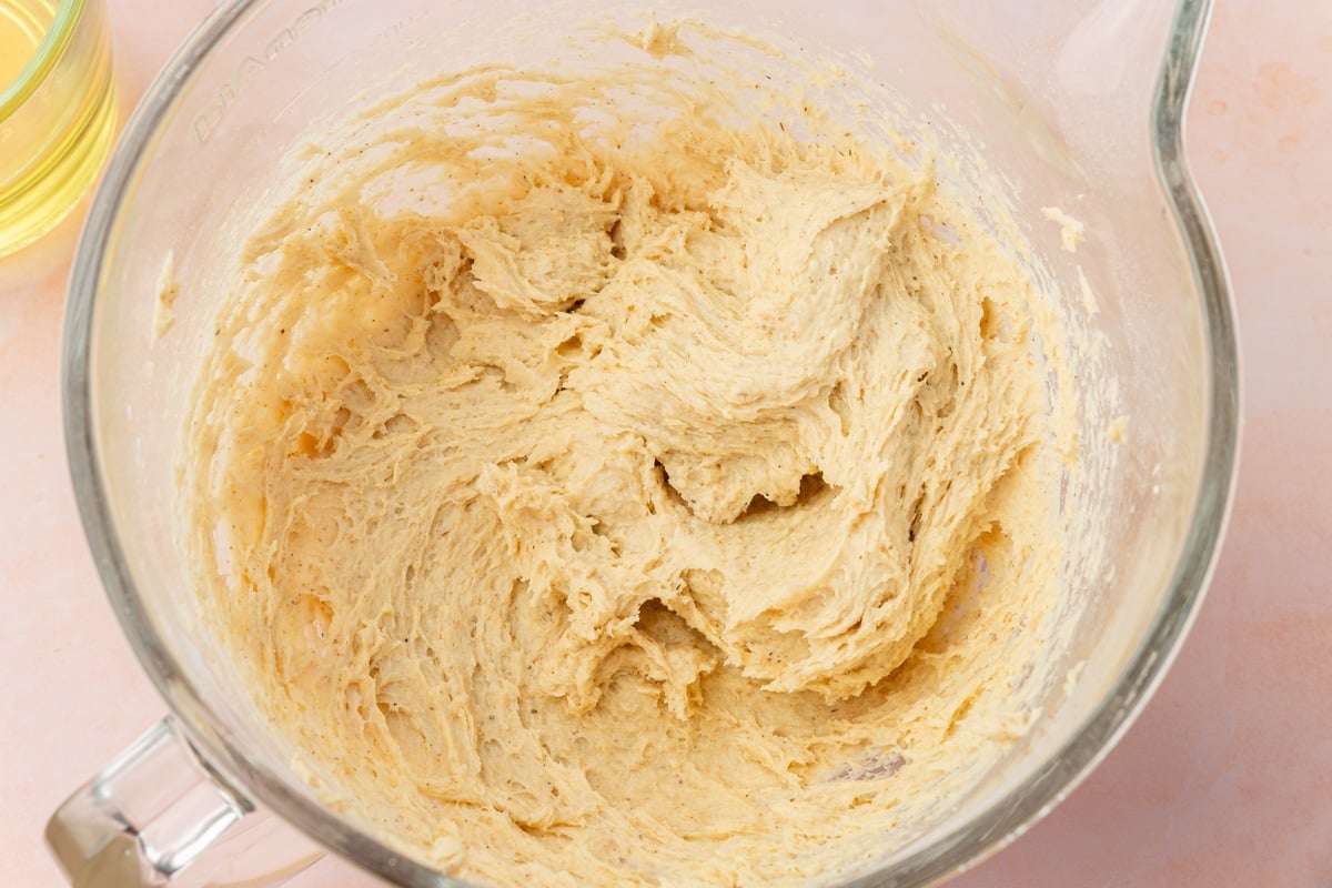 A glass mixing bowl filled with gluten-free bread dough sits on a light pink surface. The dough appears soft and well-mixed, with a few air pockets visible on its surface.