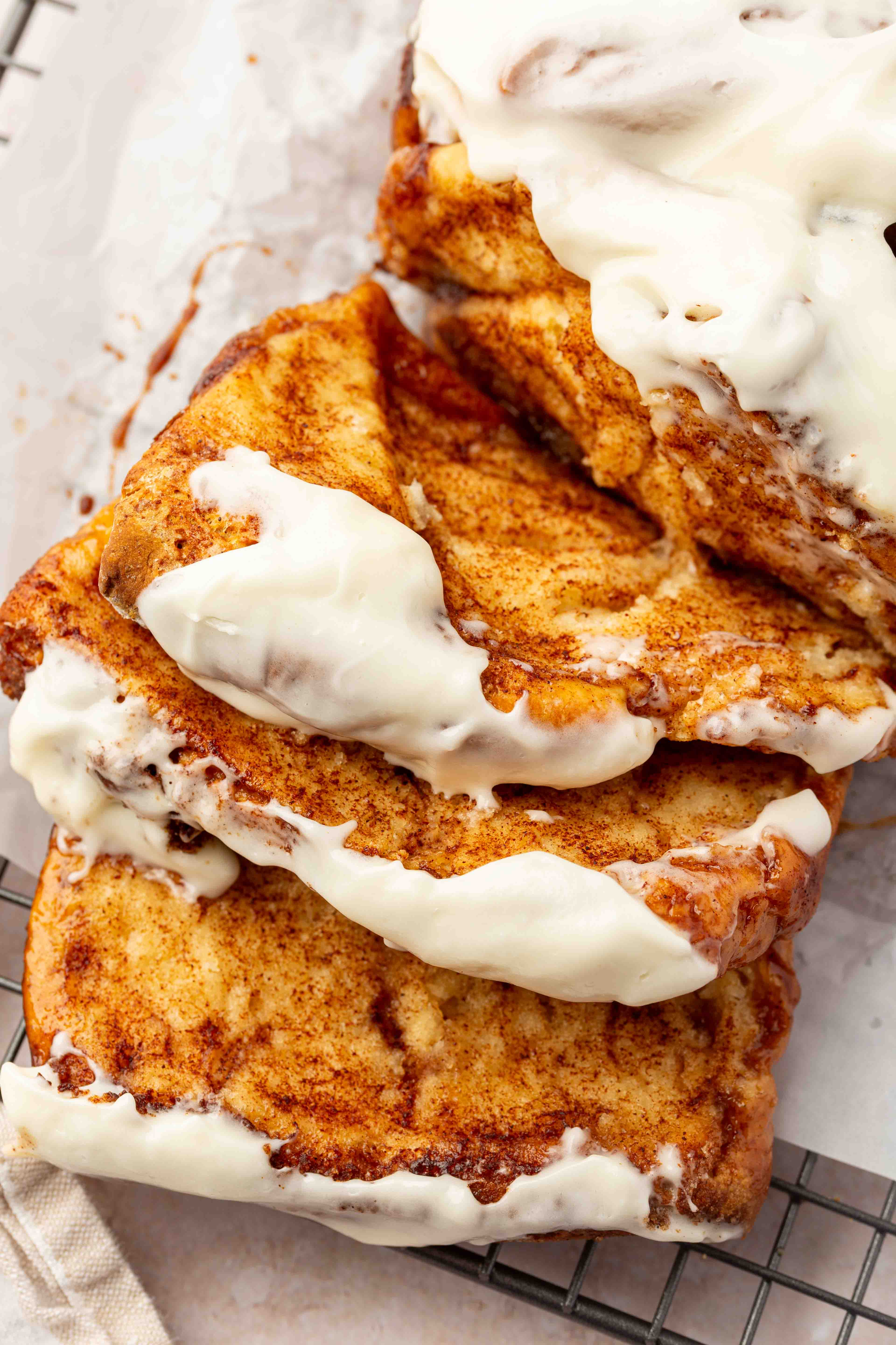 Four slices of gluten-free cinnamon bread topped with creamy white cream cheese frosting are stacked on a wire cooling rack lined with parchment paper.