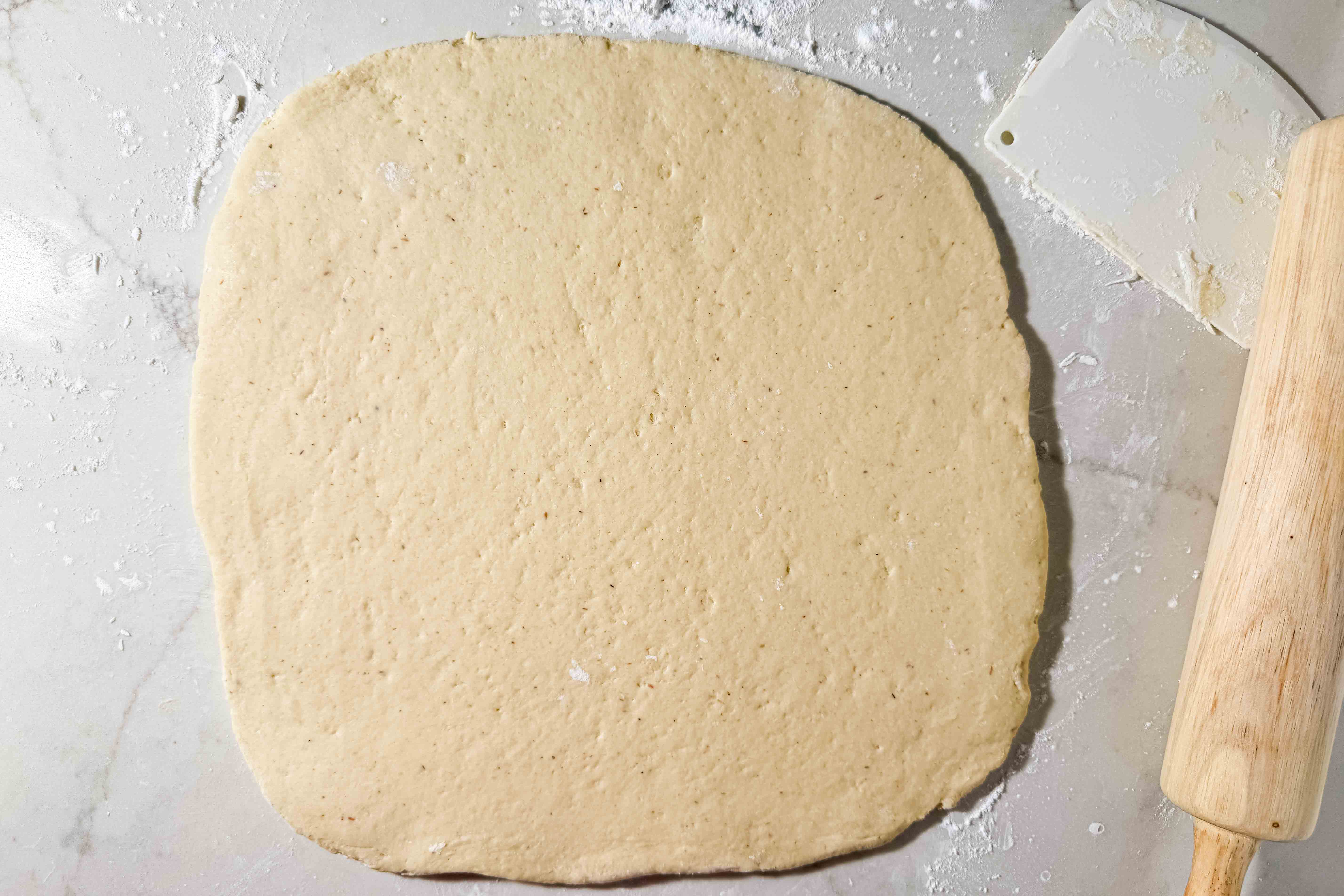 A square piece of gluten-free enriched dough is rolled out on a floured surface with a rolling pin and a dough scraper nearby.