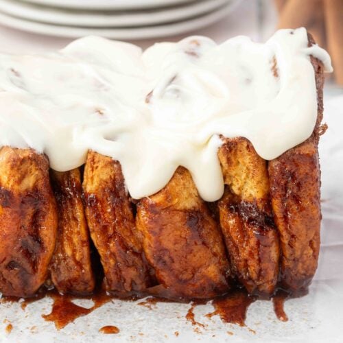 A loaf of cinnamon pull-apart bread with cream cheese icing sits on parchment paper atop a cooling rack. Stacked plates and cinnamon sticks are visible in the background.