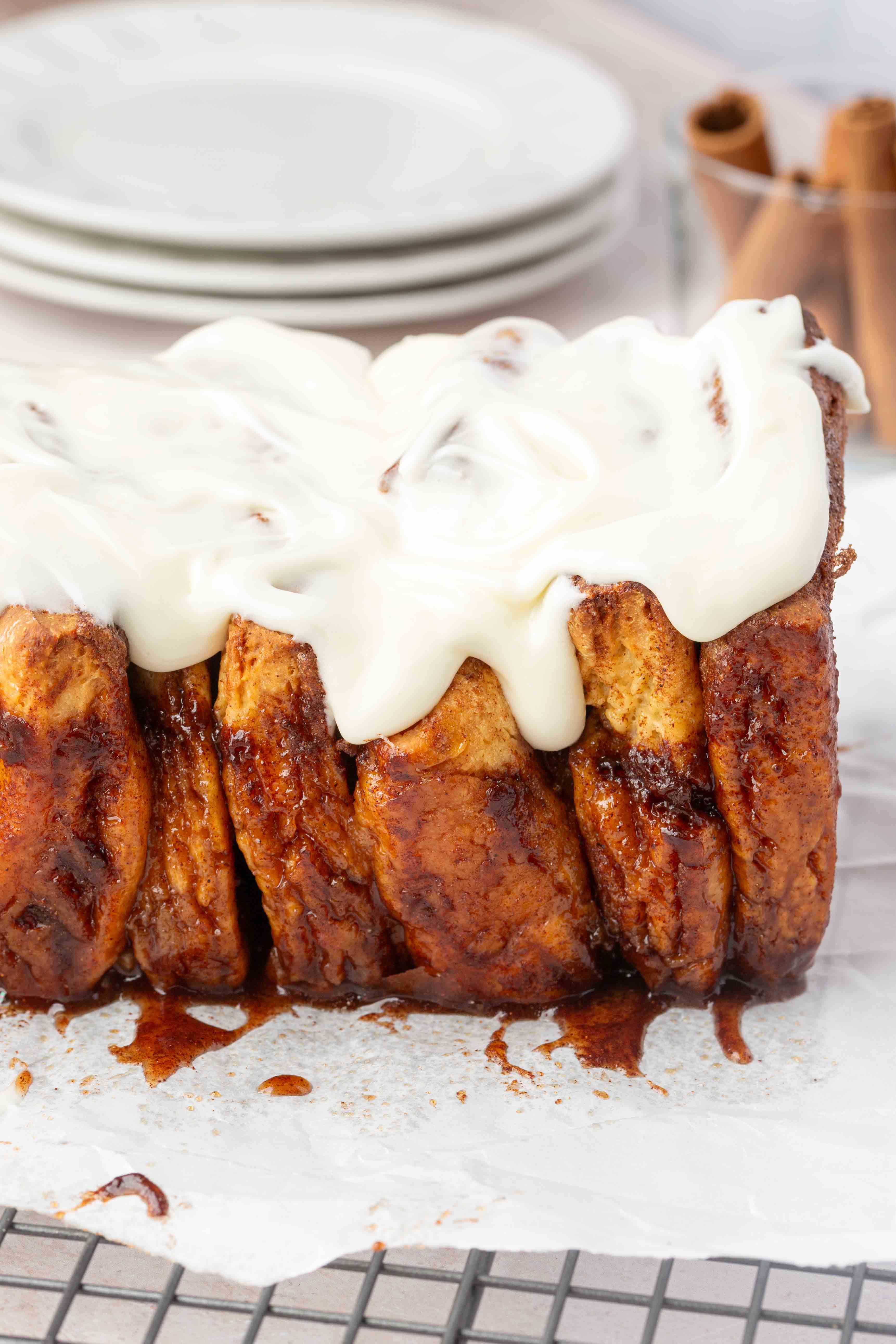 A loaf of cinnamon pull-apart bread with cream cheese icing sits on parchment paper atop a cooling rack. Stacked plates and cinnamon sticks are visible in the background.