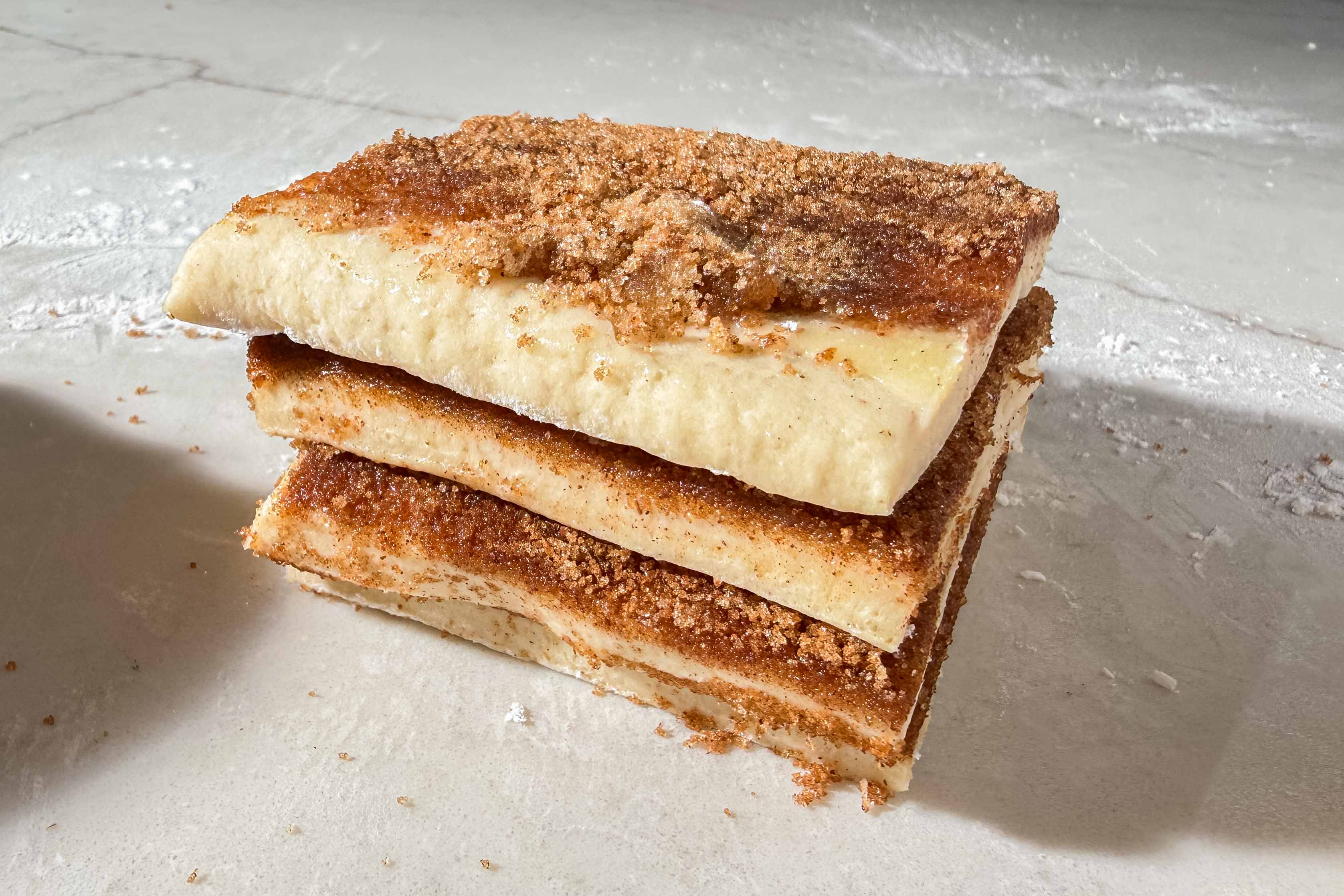 Four stacked rectangles of dough coated with cinnamon sugar, resting on a lightly floured surface, with extra cinnamon sugar sprinkled on top.