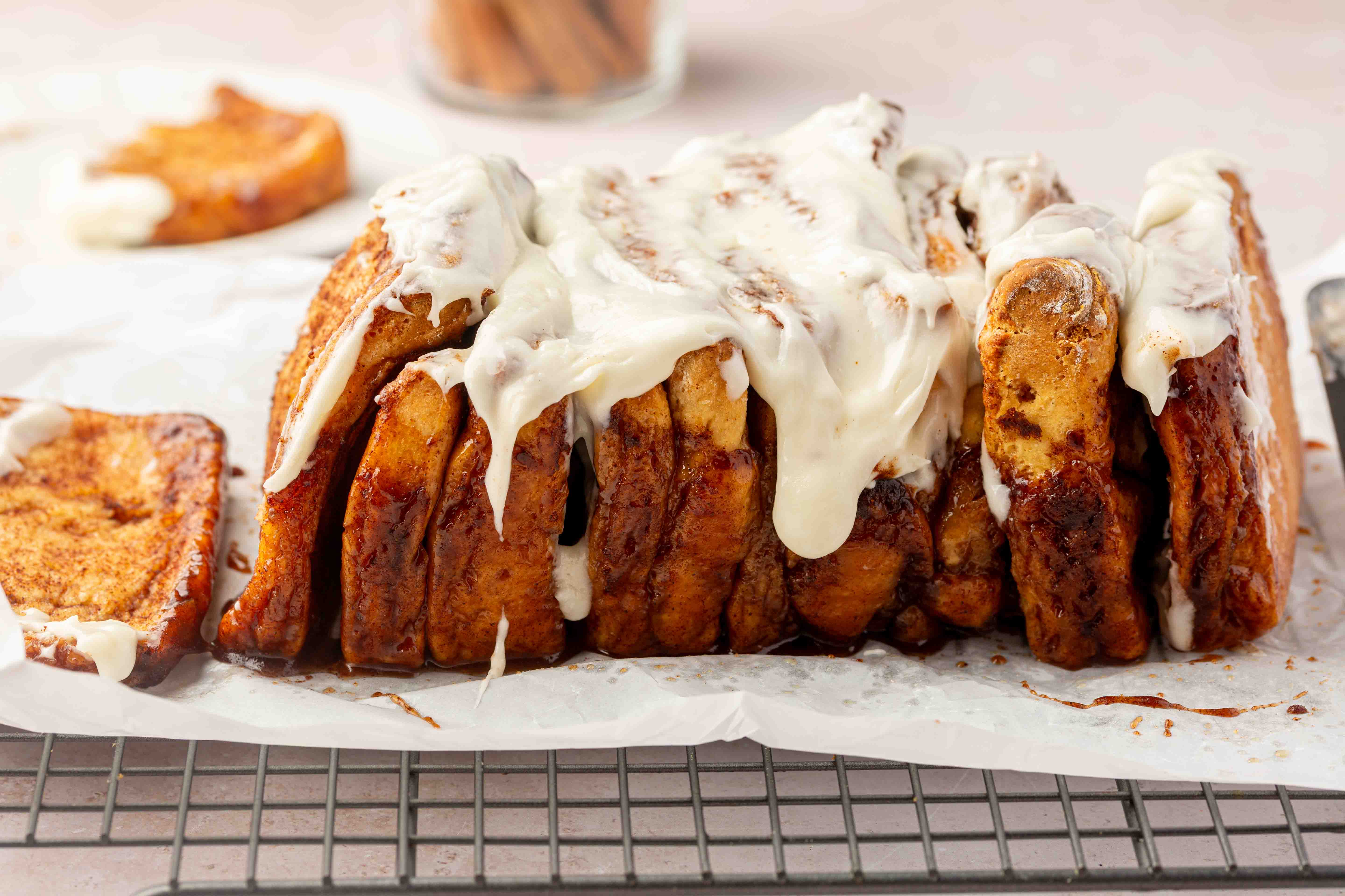 Sliced cinnamon pull-apart bread topped with white icing sits on parchment paper over a cooling rack, with one piece separated and icing dripping down the sides.