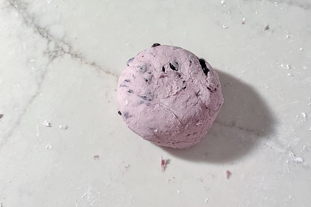 A ball of purple bagel dough with dried blueberries in it sits on a lightly floured white marble surface, casting a soft shadow to the right.