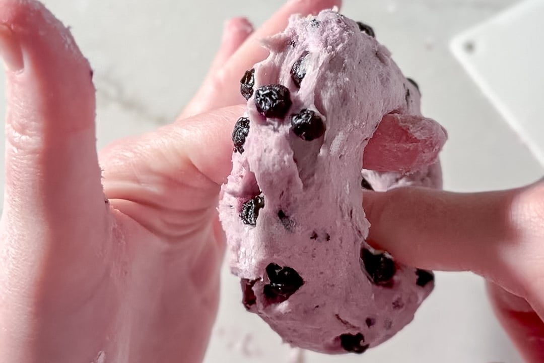 A close-up of hands stretching purple dough with embedded blueberries, with dough sticking to the fingers.