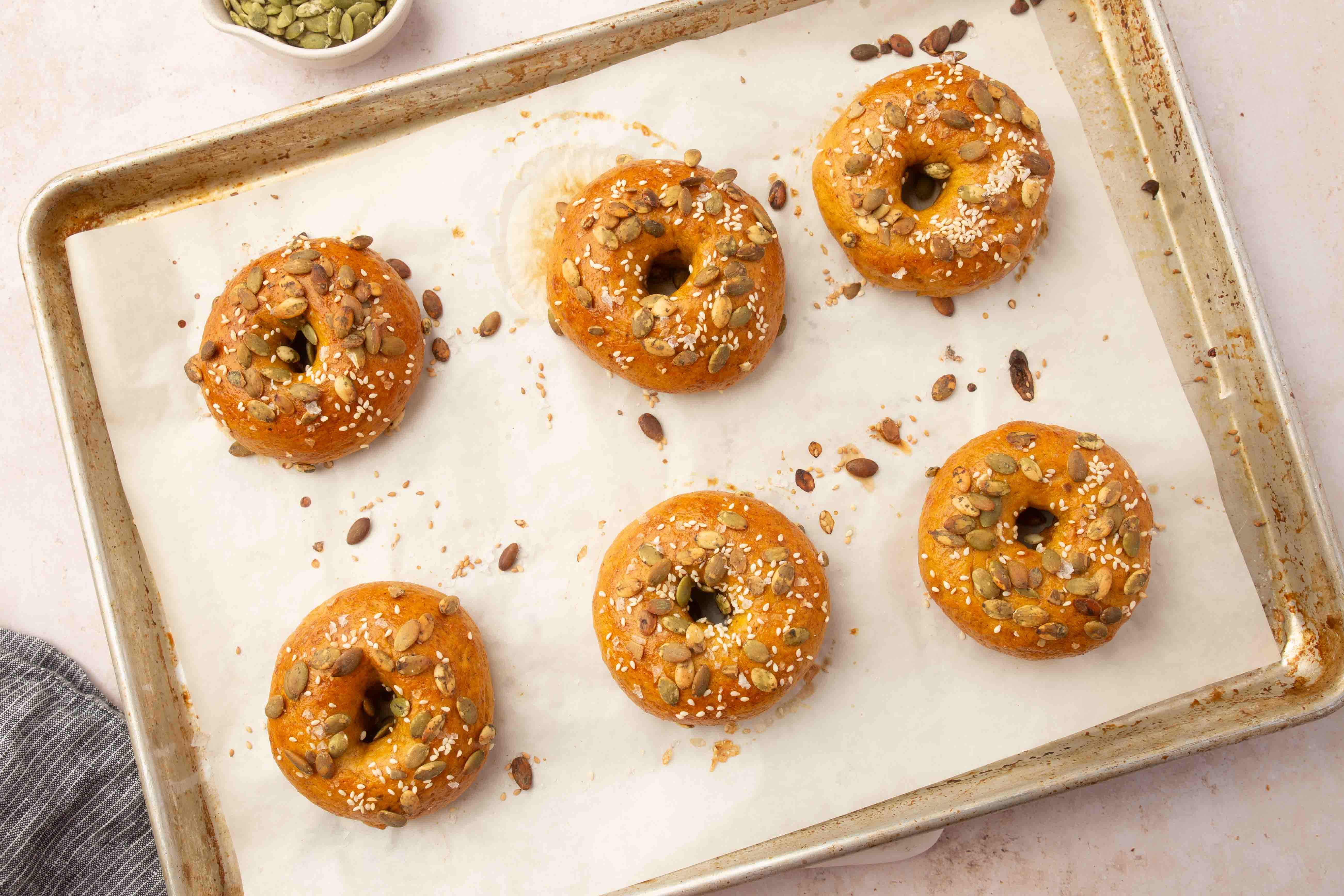 Six golden-brown bagels topped with pumpkin seeds and sesame seeds on a parchment-lined baking sheet.