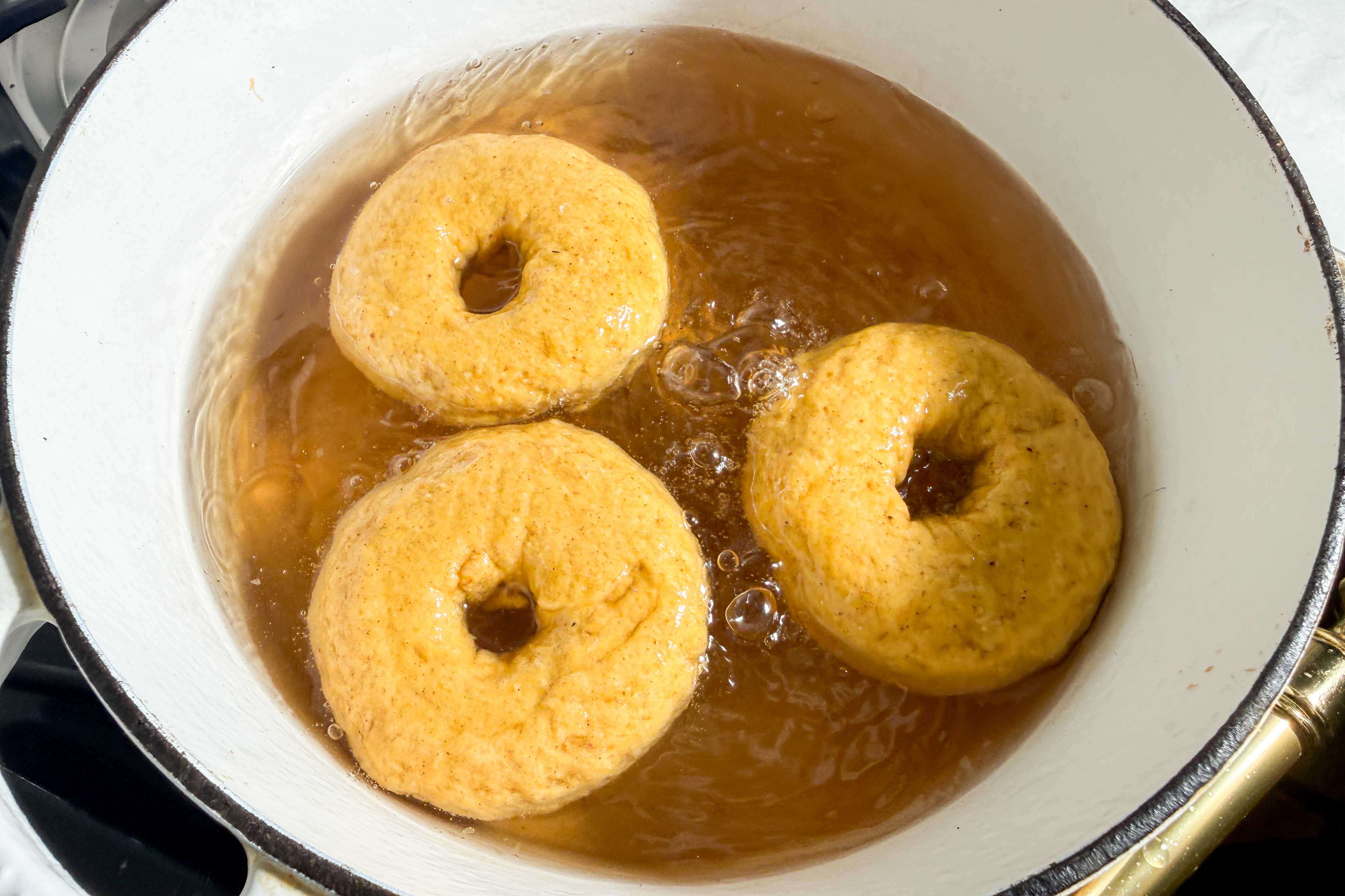 Three pumpkin bagels boiling in a dutch oven filled with water and brown sugar.