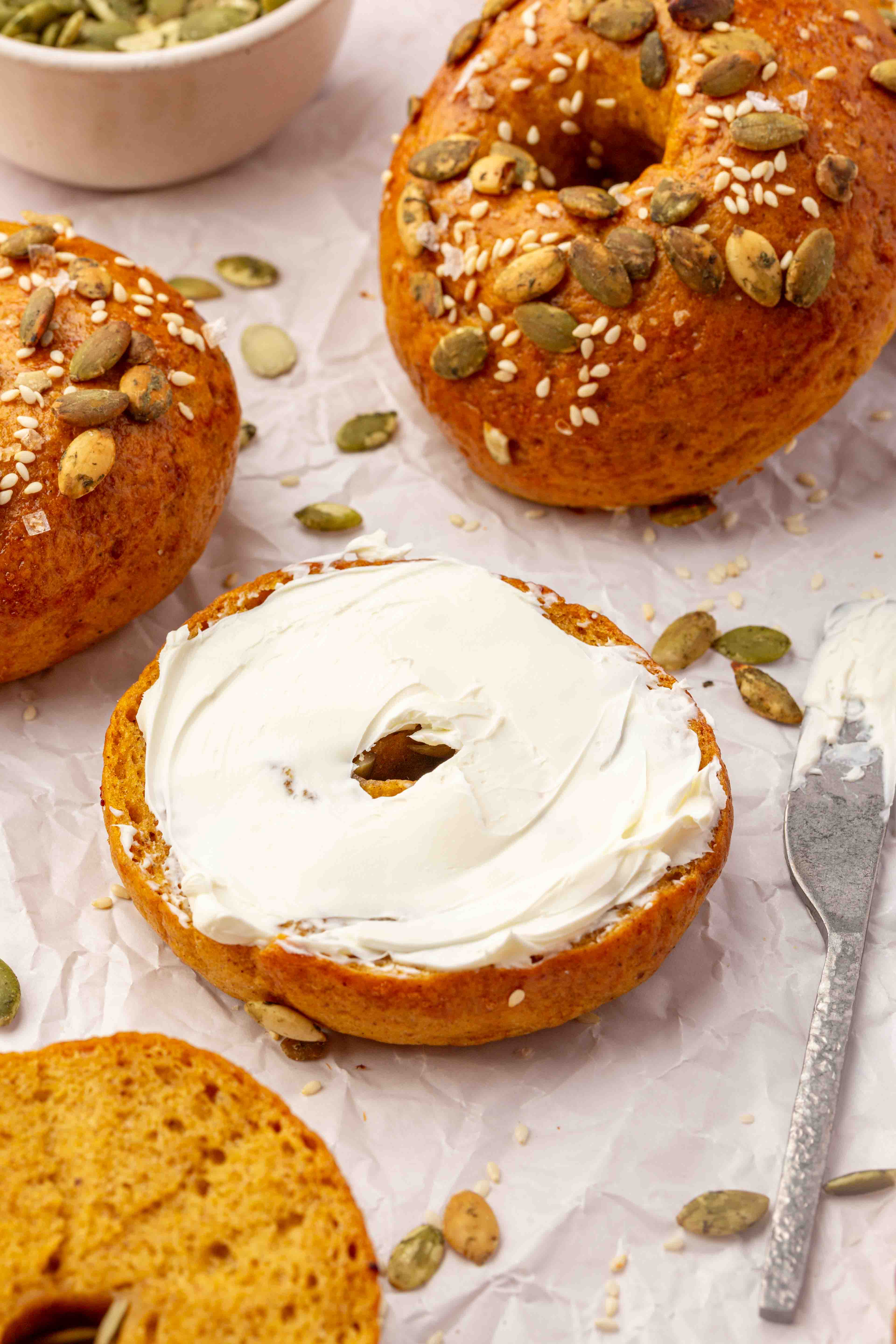 Pumpkin bagels topped with sesame seeds and pumpkin seeds, one spread with cream cheese next to a spreading knife.