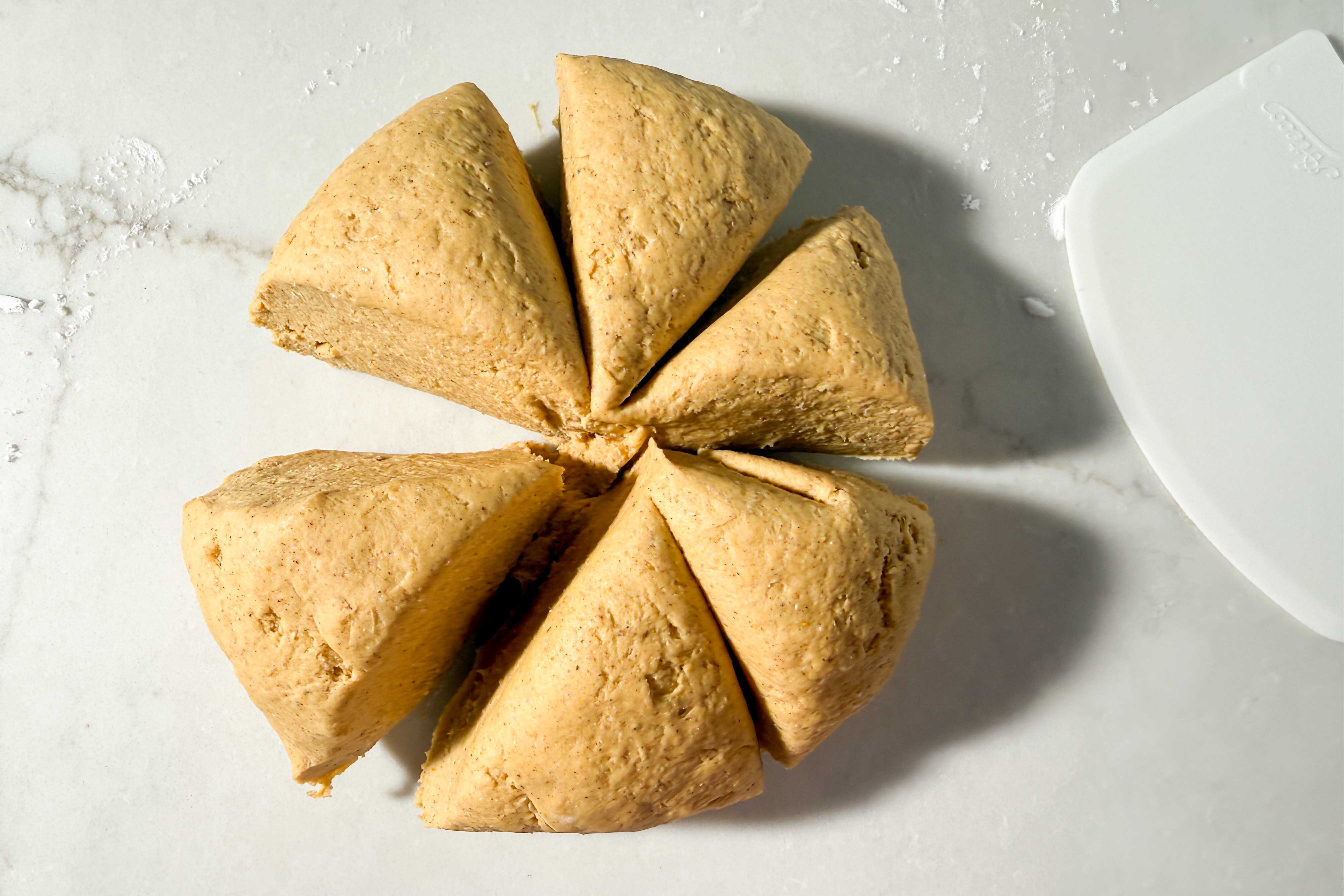 Six triangular portions of raw gluten-free pumpkin bagel dough arranged in a circle on a countertop next to a plastic dough scraper.