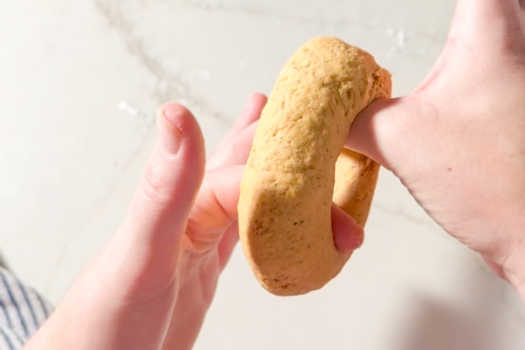Hands forming a ring of unbaked bagel dough.