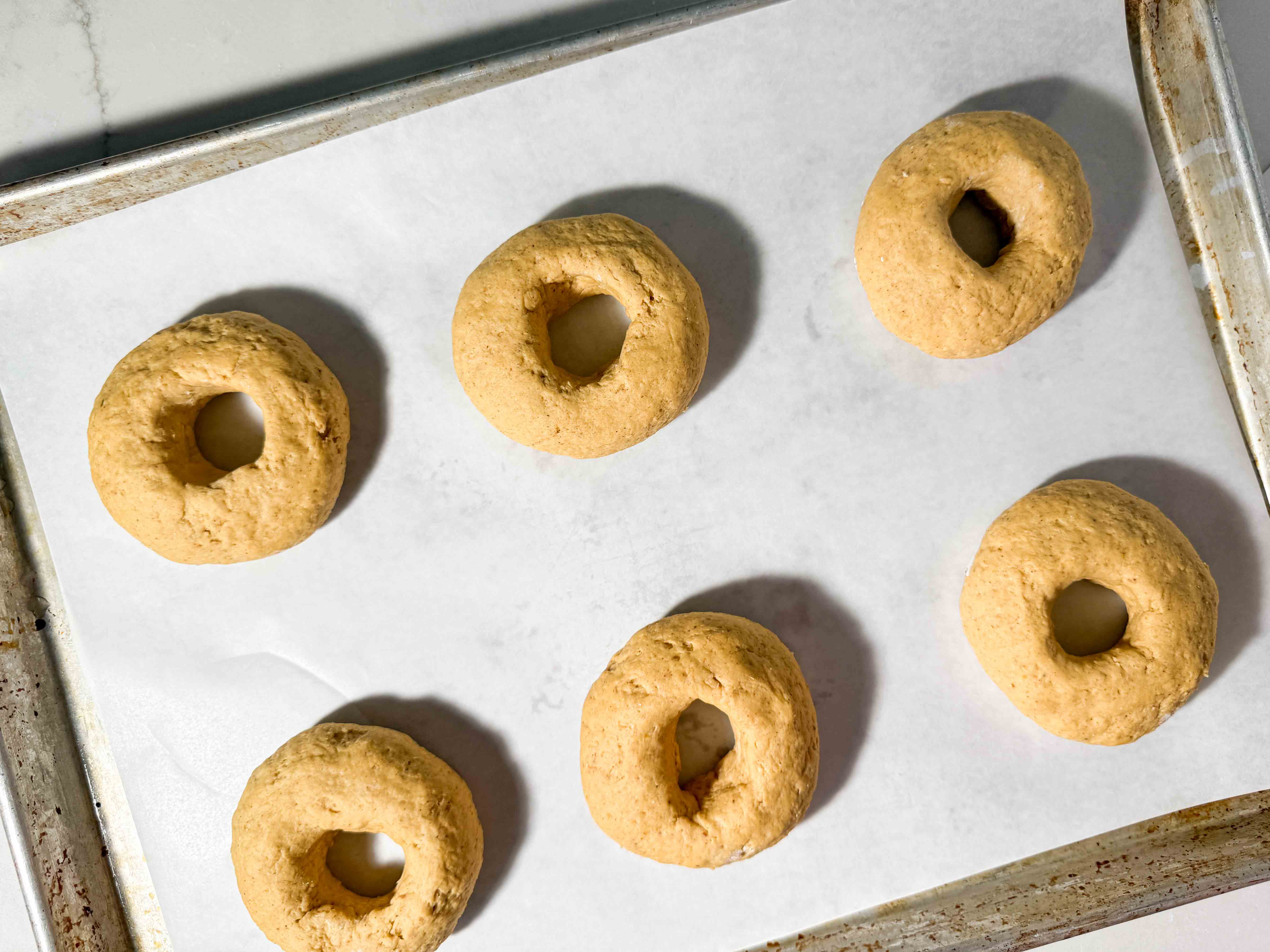 Six raw, pumpkin bagel dough rounds arranged on a parchment-lined baking sheet before proofing.
