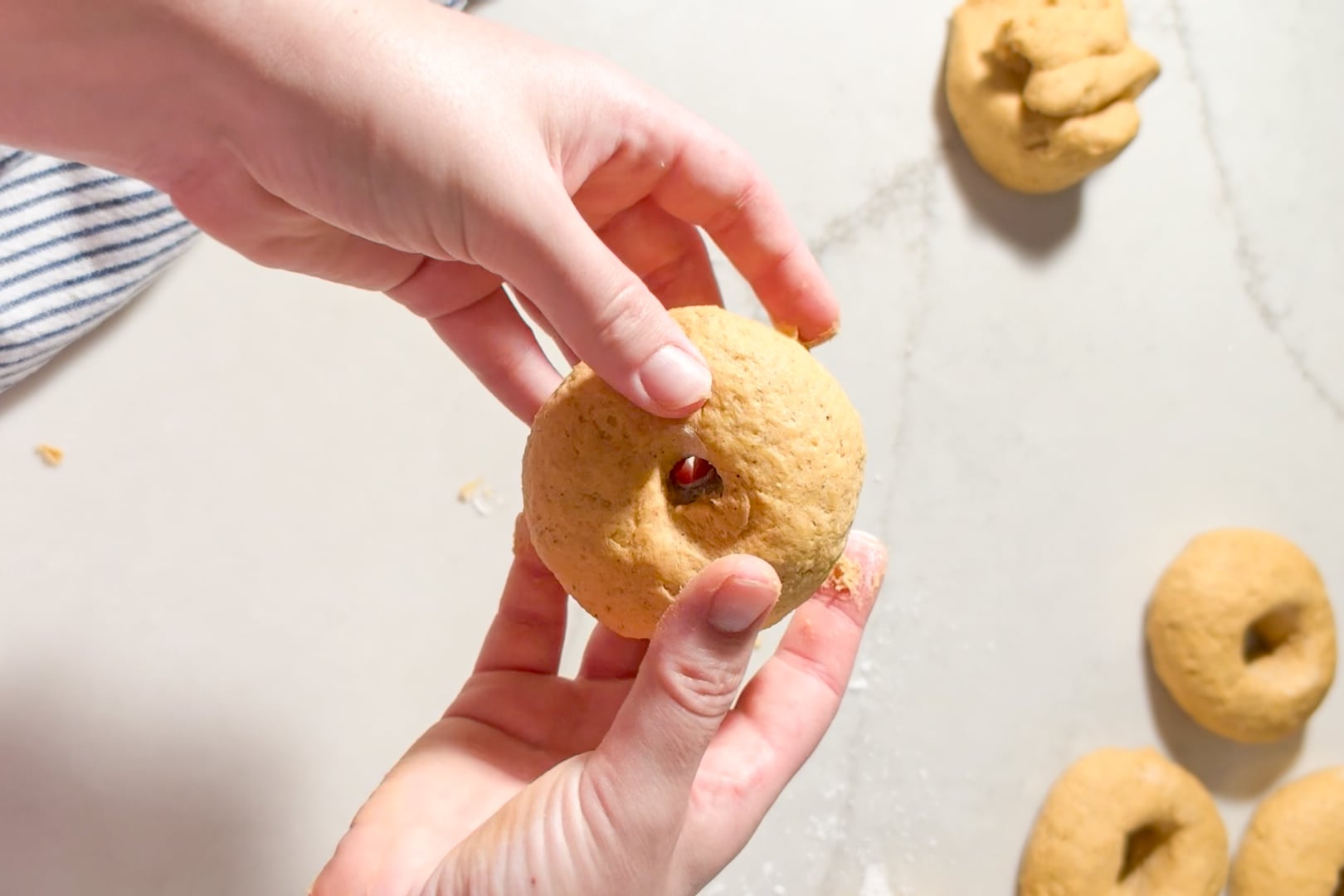 Hands shaping a piece of dough into a bagel shape on a countertop with other shaped dough pieces nearby.