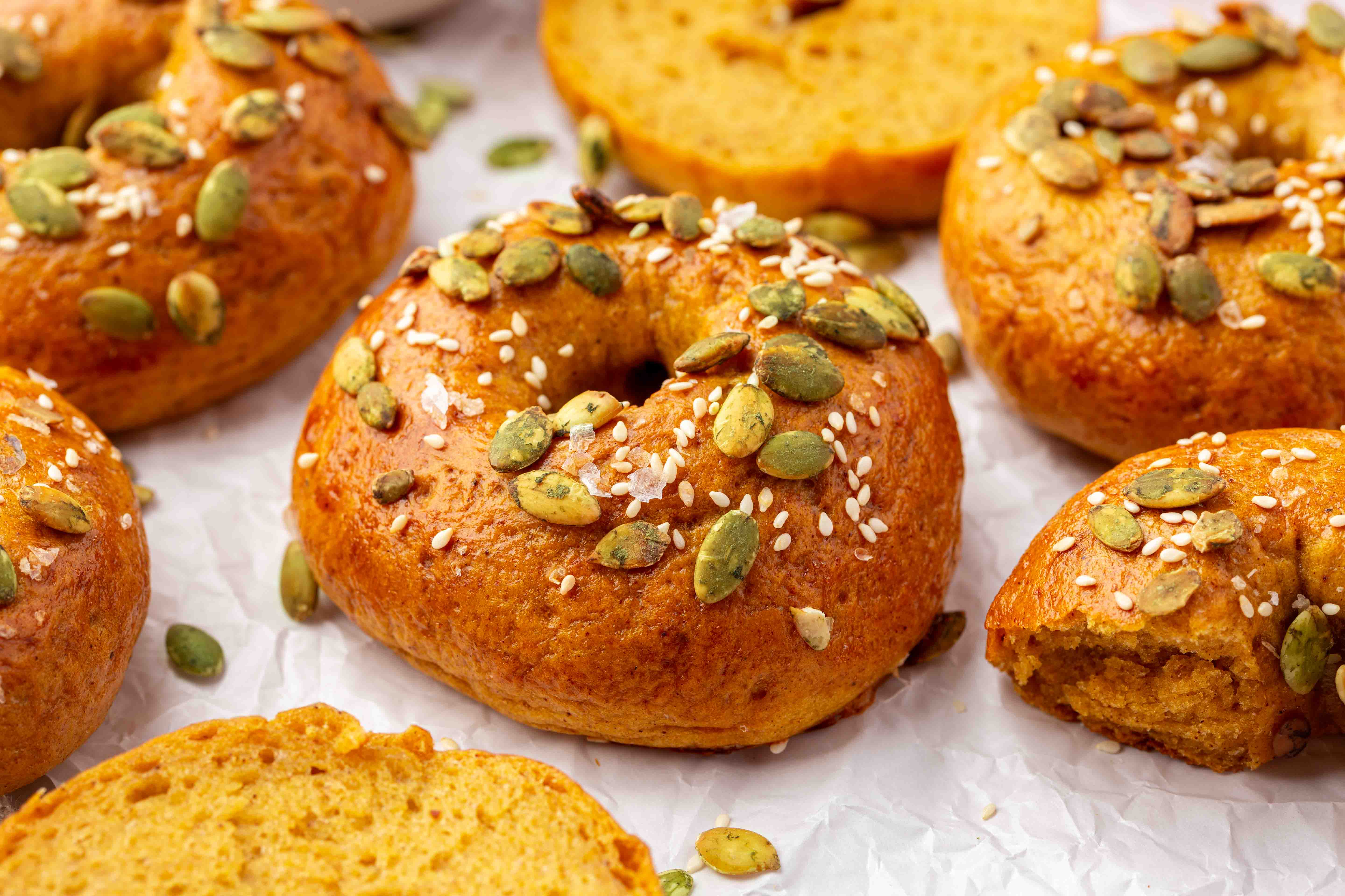 Several gluten-free pumpkin bagels topped with green pumpkin seeds, sesame seeds, and coarse salt, resting on crumpled parchment paper.