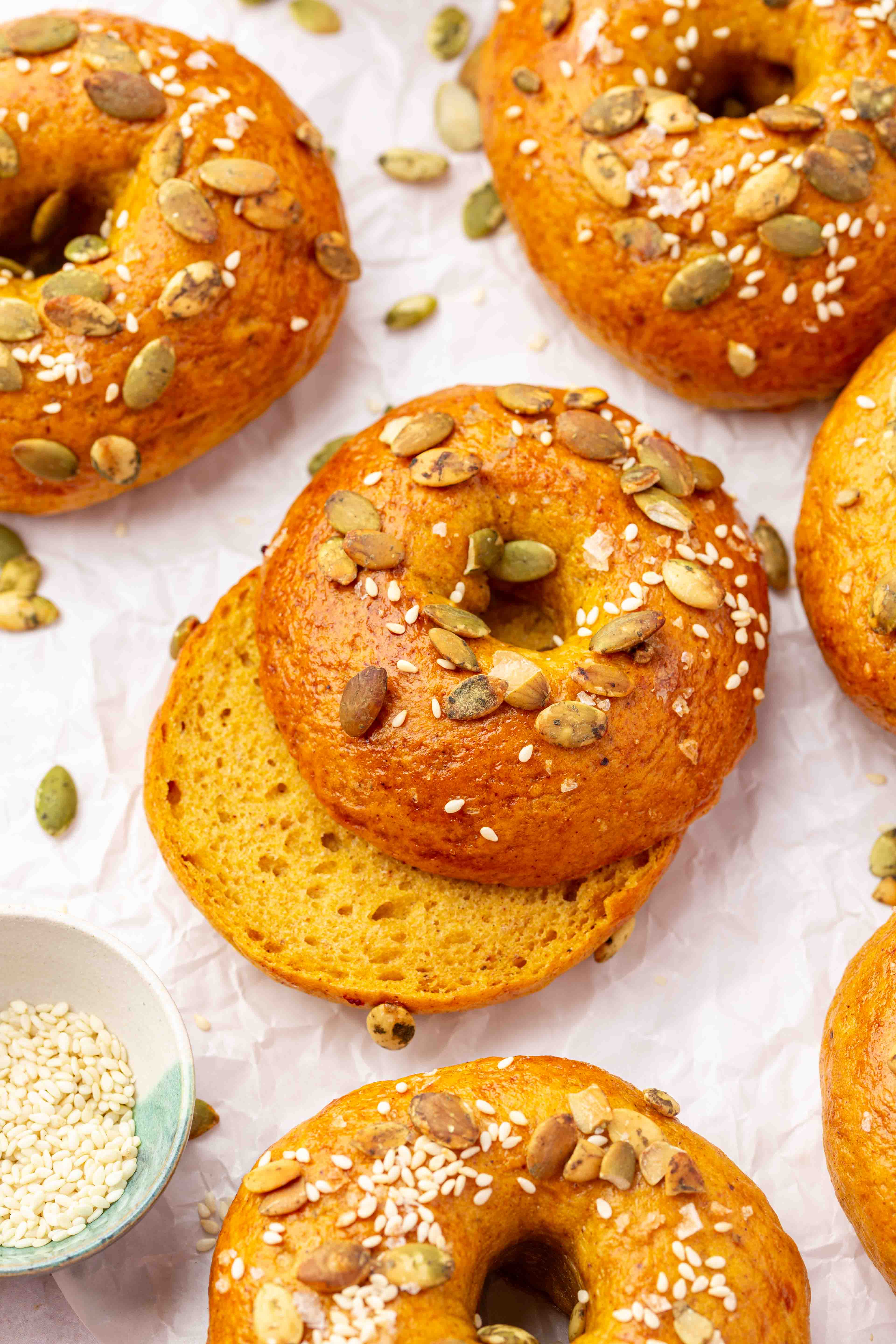 Golden pumpkin bagels topped with pumpkin seeds and sesame seeds on parchment paper, with one bagel sliced open.