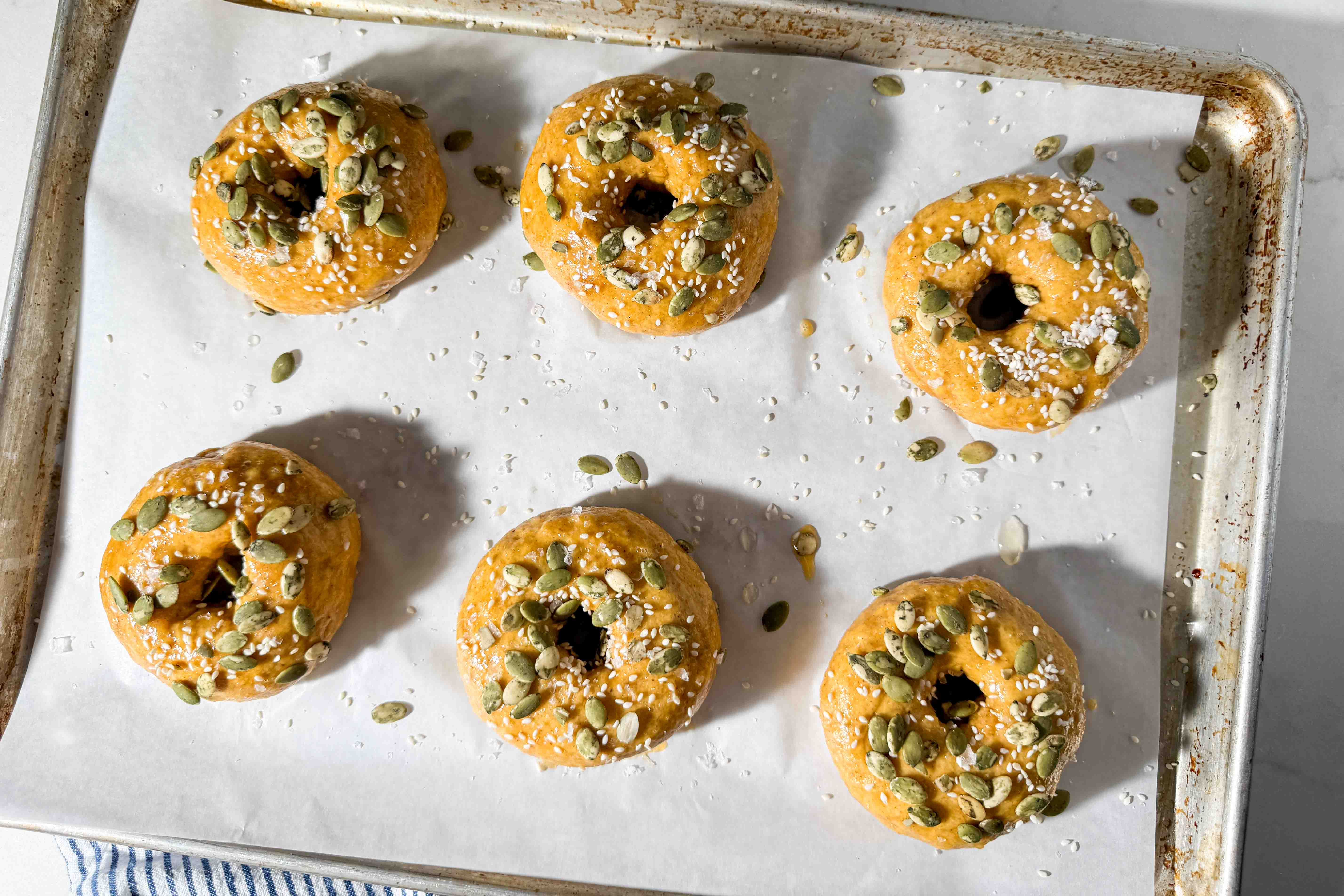 Six uncooked pumpkin bagels topped with pumpkin seeds and sesame seeds arranged on a parchment-lined baking sheet.