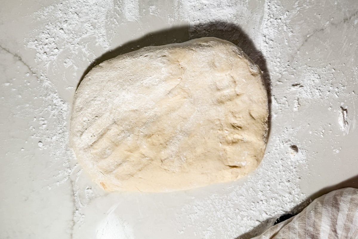 A flattened piece of gluten-free dough dusted with flour on a marble surface.
