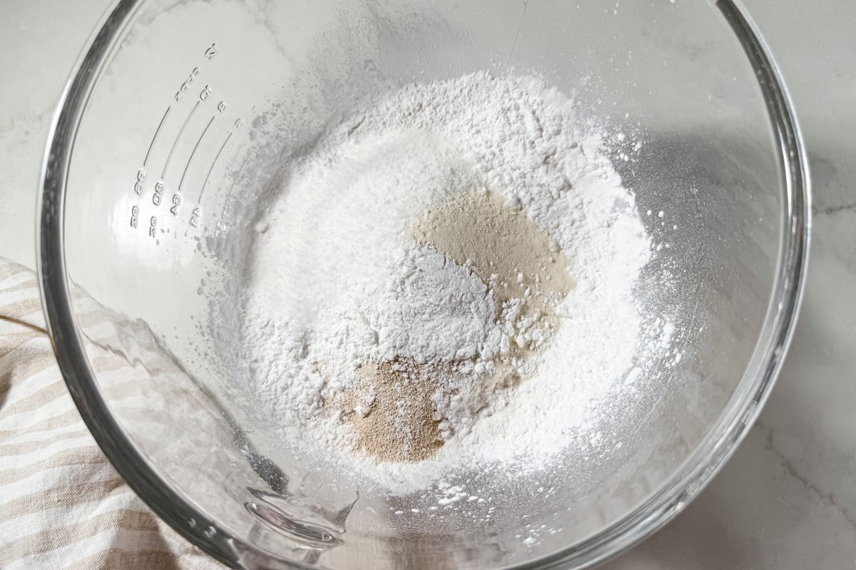 A clear glass mixing bowl containing gluten-free flour, sugar, and yeast.