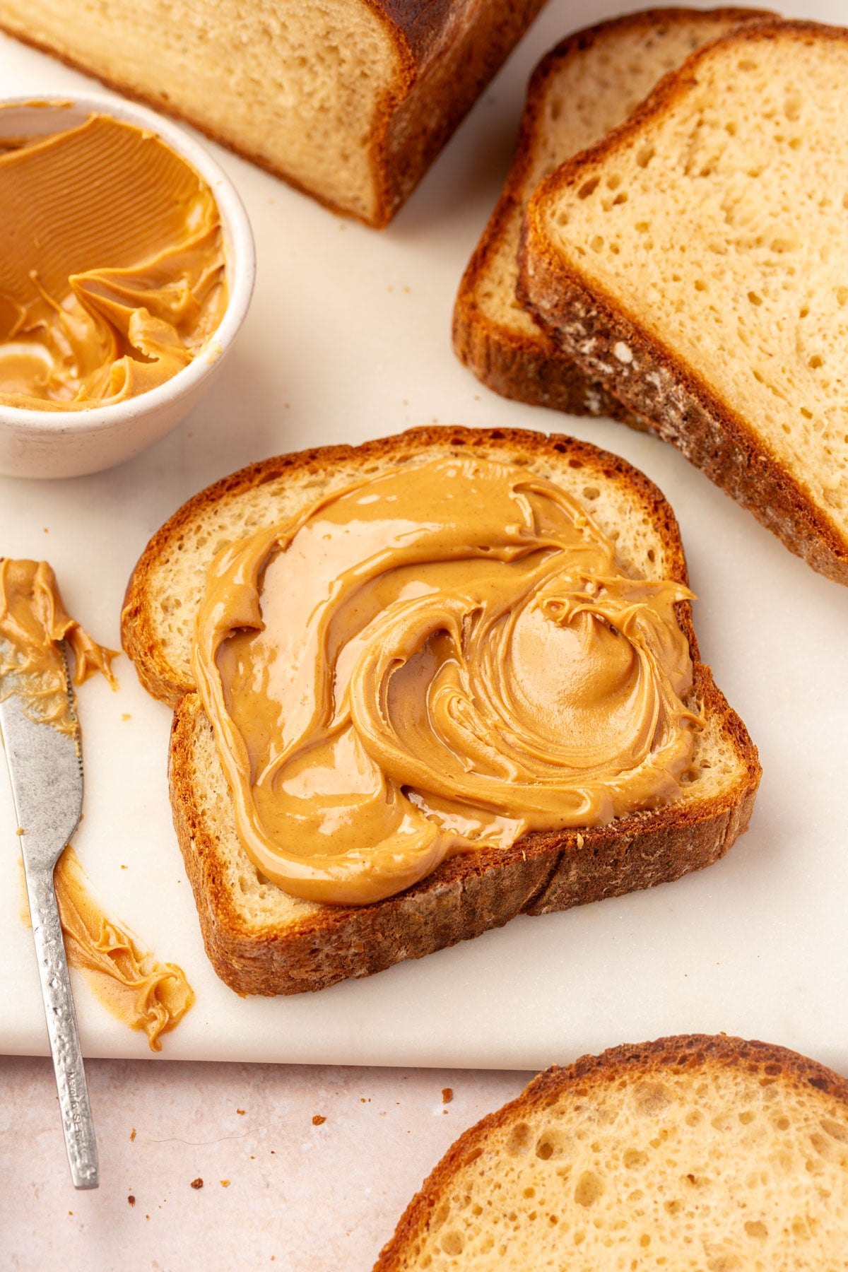 A slice of gluten-free bread spread with creamy peanut butter next to a bowl of peanut butter, a butter knife, and additional slices of bread.