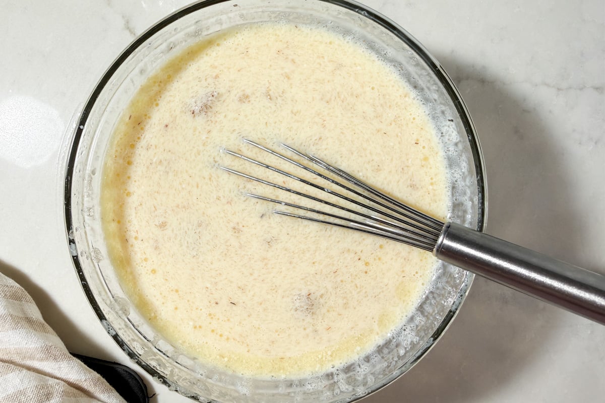 A wire whisk resting in a glass bowl filled with a milk, egg, and psyllium gel mixture.